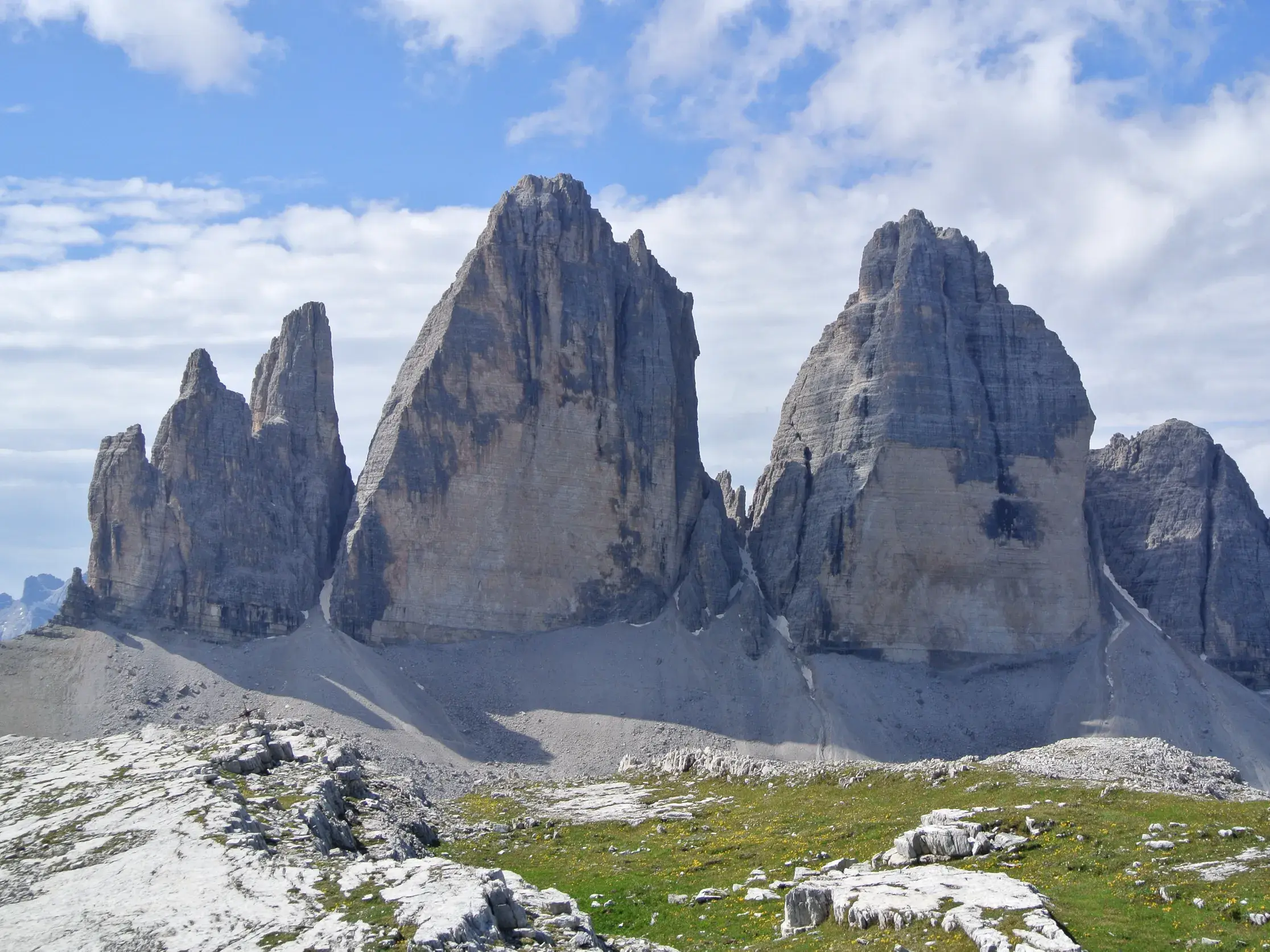 Blick von einer Almwiese auf die markanten Felsgipfel der Drei Zinnen in den Sextener Dolomiten unter blauem Himmel.