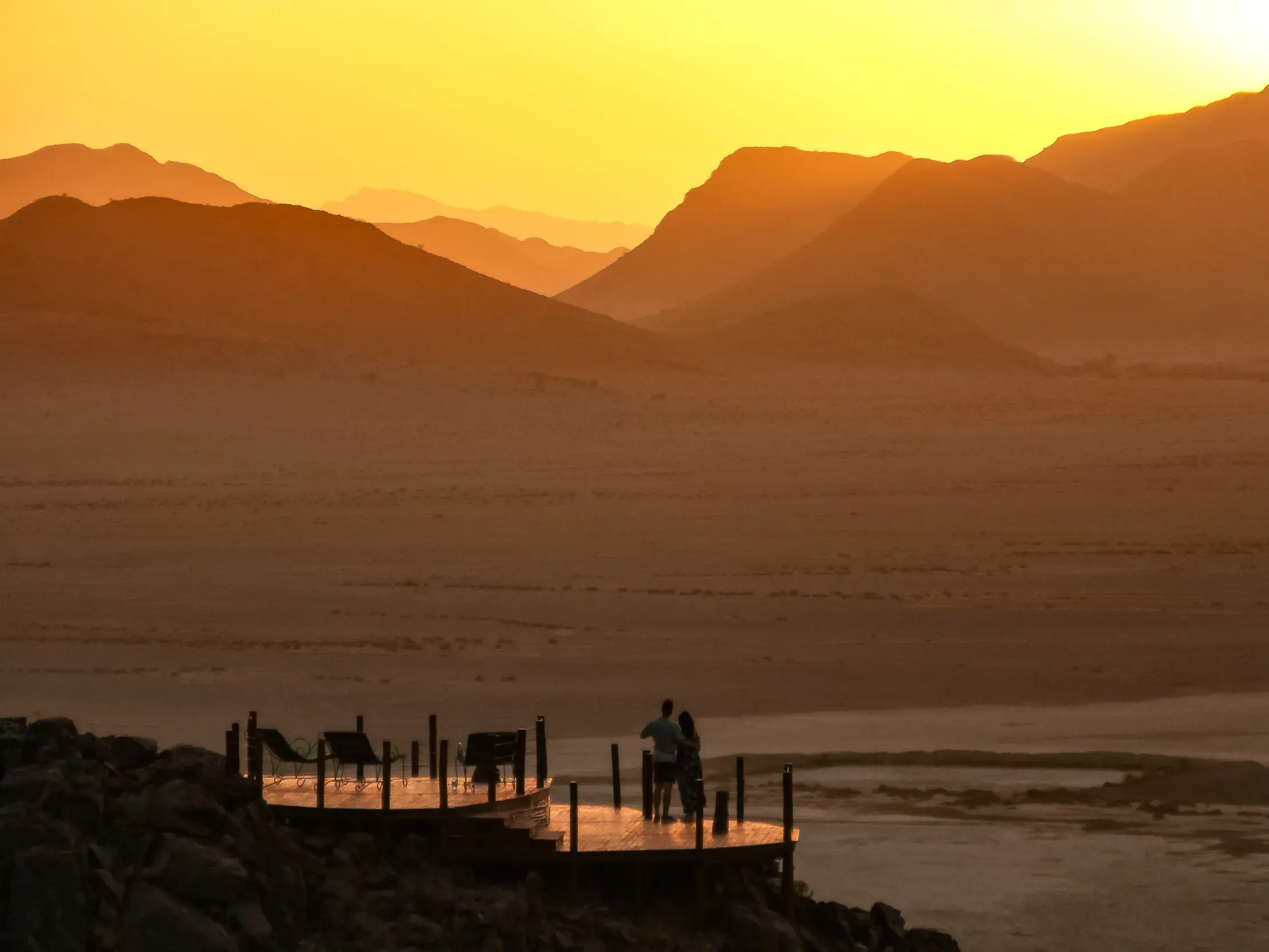 Paar genießt den Sundowner auf einer Lodge-Terrasse mit Blick auf die weite Berglandschaft der Wüste Namibias bei Sonnenuntergang.