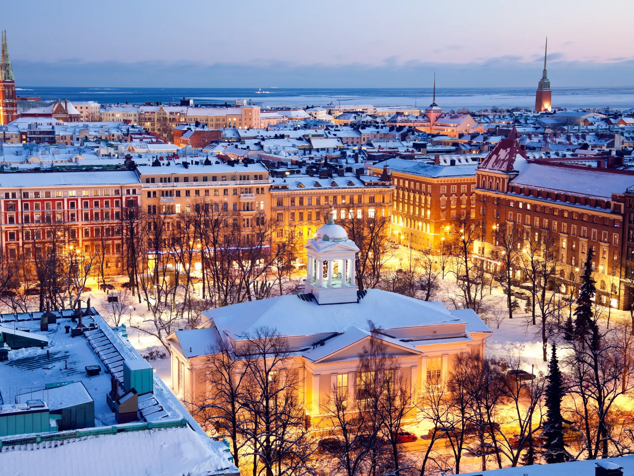 Verschneite Dächer von Helsinki in der Abenddämmerung mit beleuchteten Gebäuden und Blick auf die winterliche Ostsee.