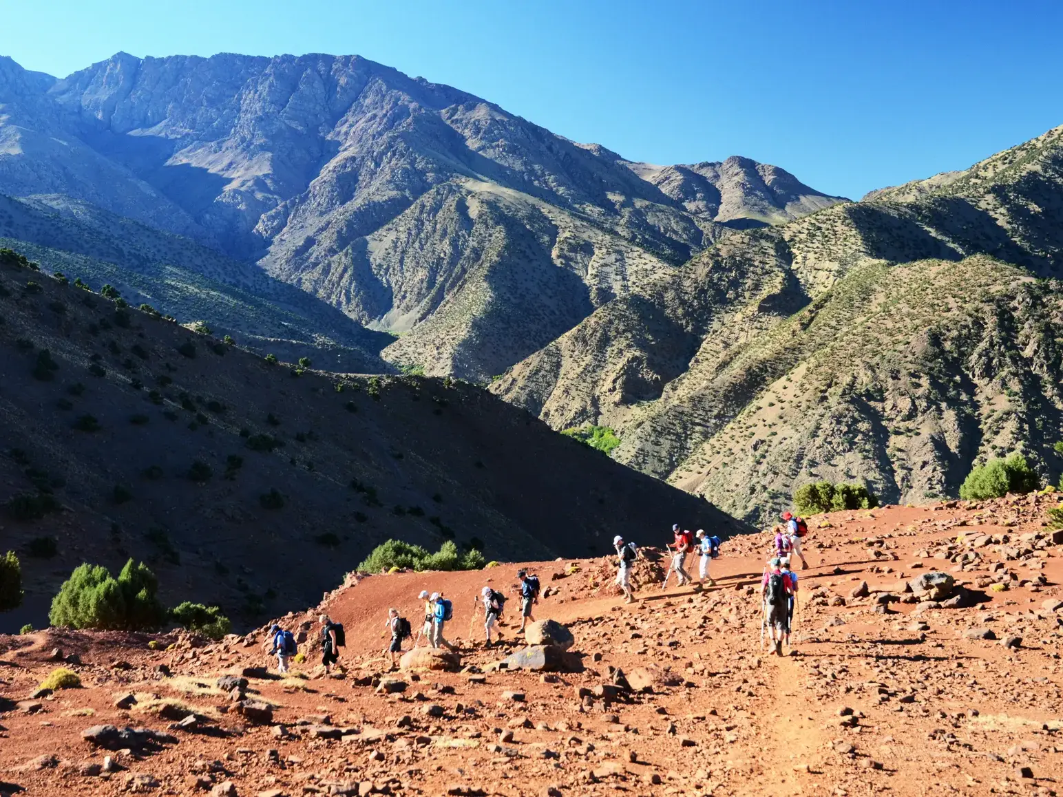 Eine Trekkinggruppe wandert auf einem schmalen Pfad über rote, steinige Erde im Hohen Atlas in Marokko.