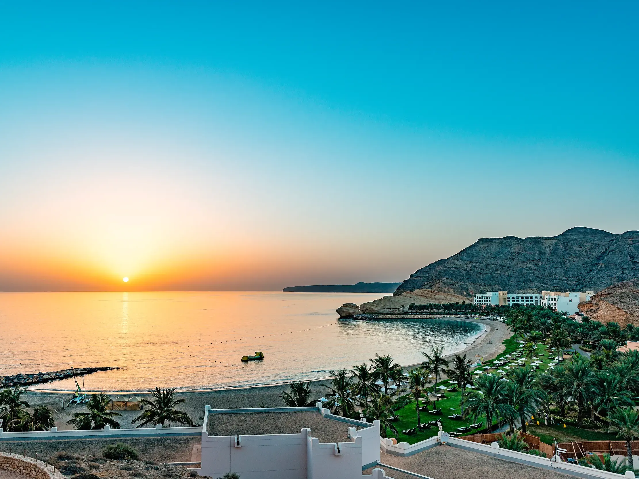 Malerischer Sonnenaufgang über einer Bucht an der Küste von Salalah im Oman mit Sandstrand, Palmen und Hotelanlage am Fuße der Berge.