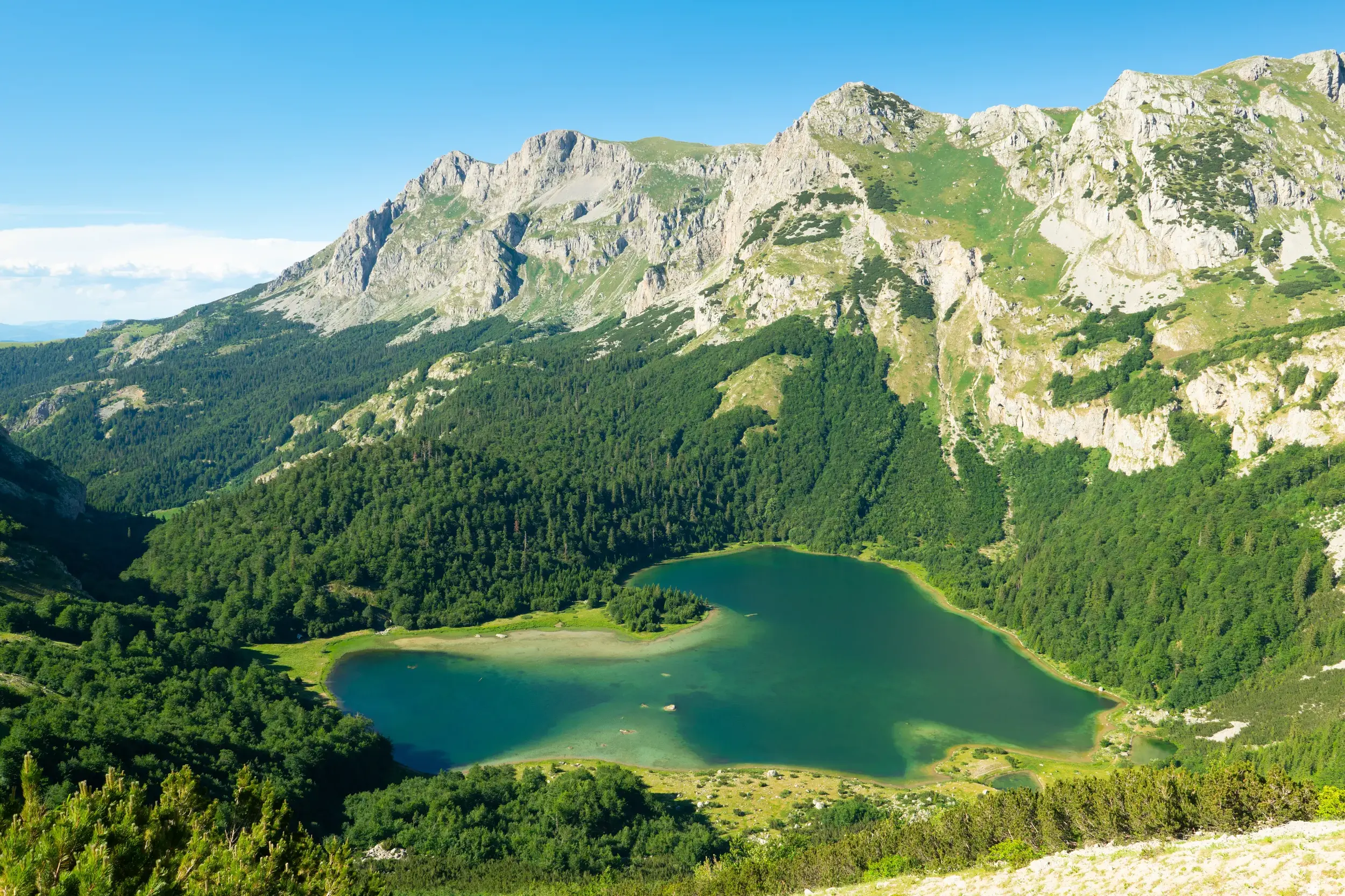 Panoramablick beim Trekking auf einen türkisen, herzförmigen Bergsee, umrahmt von dichten Wäldern und einer schroffen Gebirgskette.