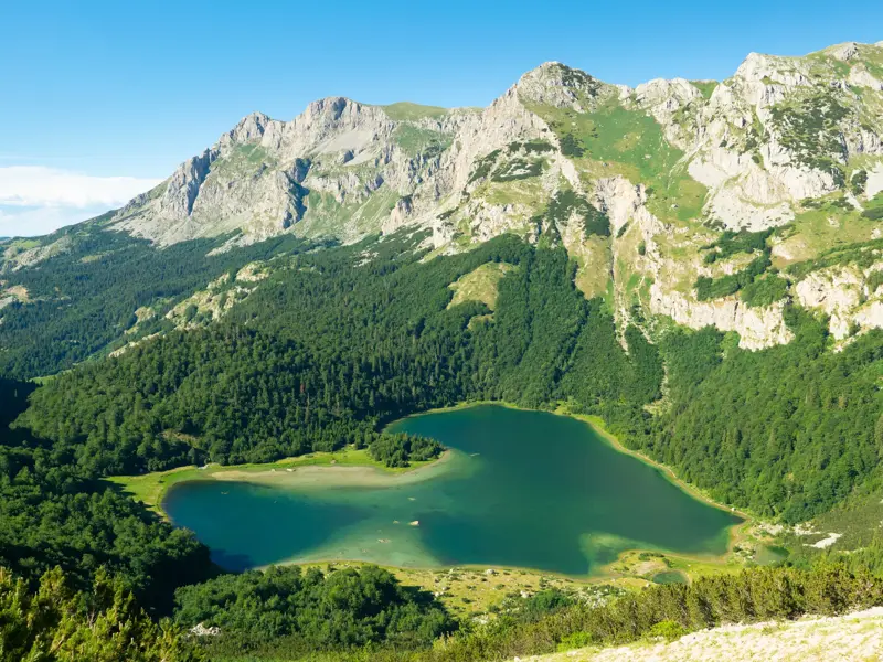 Panoramablick beim Trekking auf einen türkisen, herzförmigen Bergsee, umrahmt von dichten Wäldern und einer schroffen Gebirgskette.