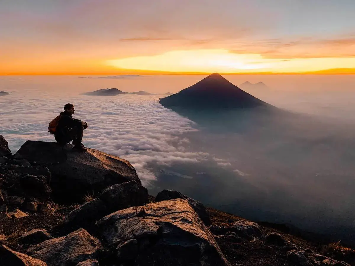 Ein Trekker sitzt auf einem Felsen am Gipfel des Vulkans Acatenango und blickt bei Sonnenaufgang über ein Wolkenmeer in Guatemala.