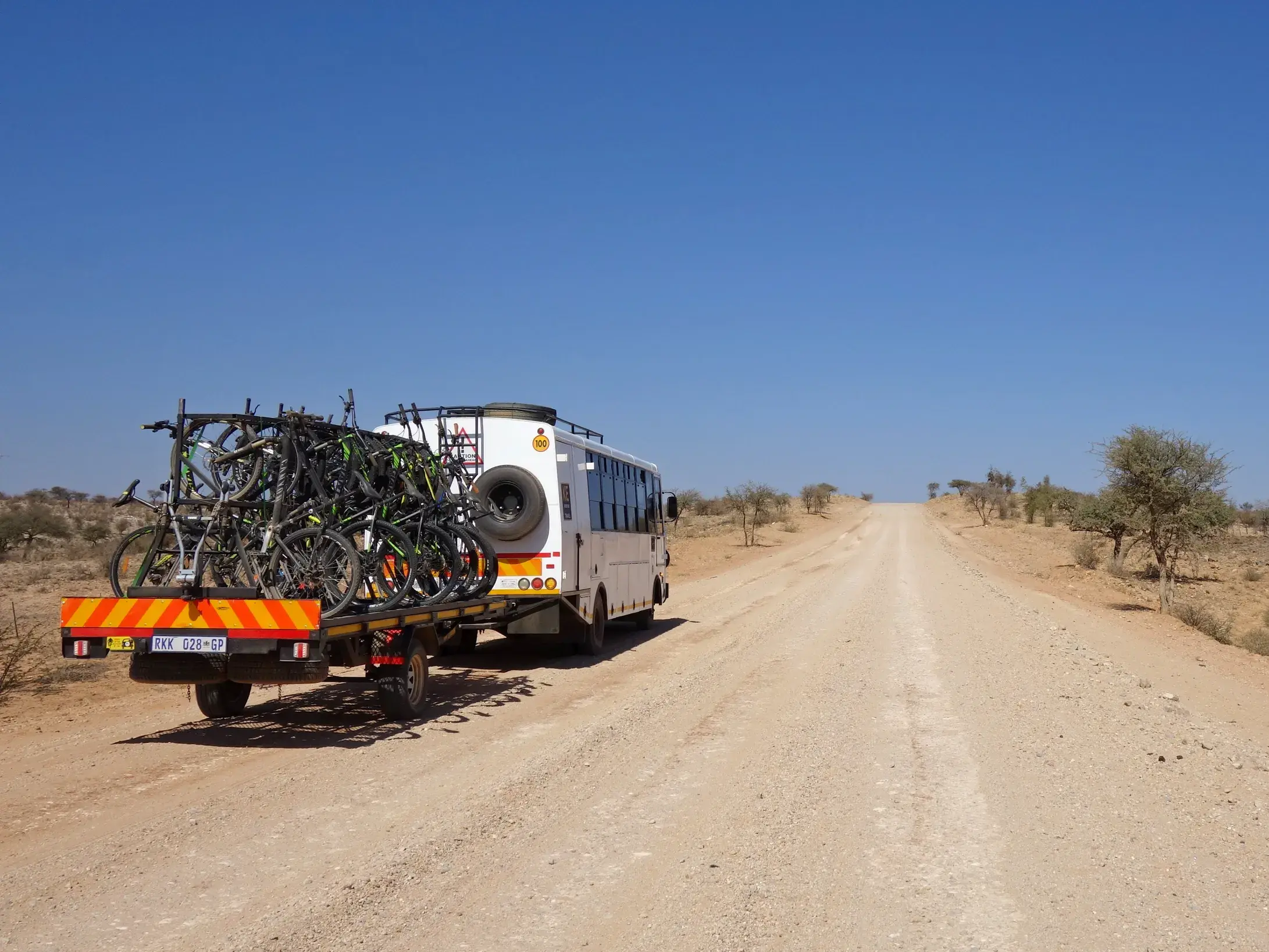 Ein weißes Begleitfahrzeug transportiert zahlreiche Mountainbikes auf einem Anhänger über eine lange Schotterpiste durch die Kalahari-Wüste.
