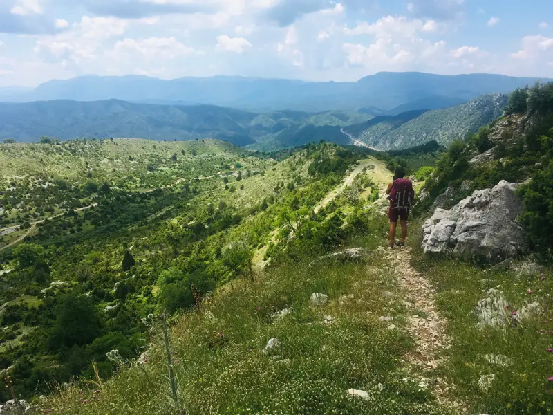 Ein Wanderer mit Rucksack auf einem Bergpfad blickt über die weite, grüne Hügellandschaft der Zagori-Region in Griechenland.