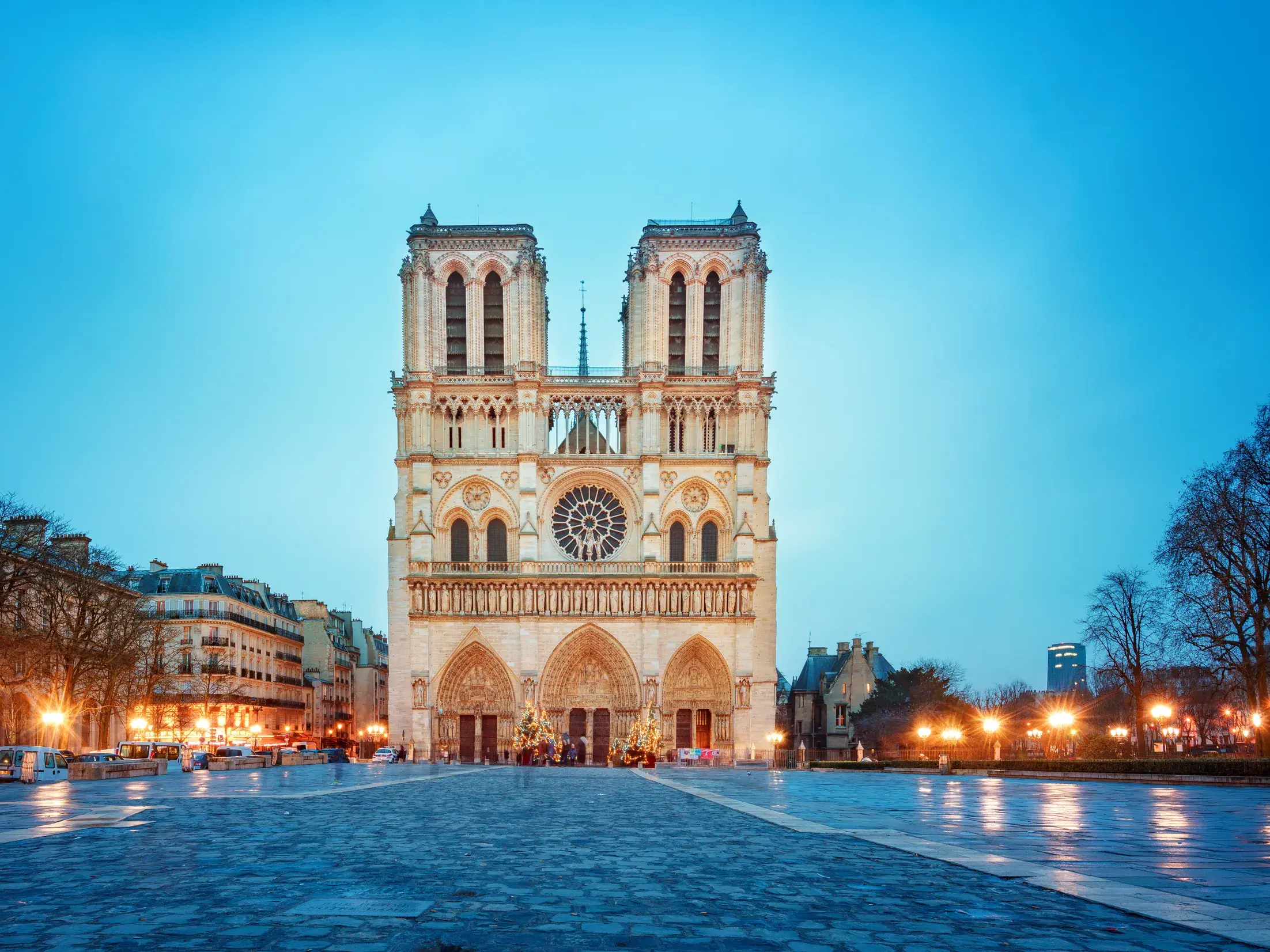 Die berühmte Kathedrale Notre-Dame in Paris bei Dämmerung. Die Lichter der Fassade spiegeln sich auf dem nassen Vorplatz.