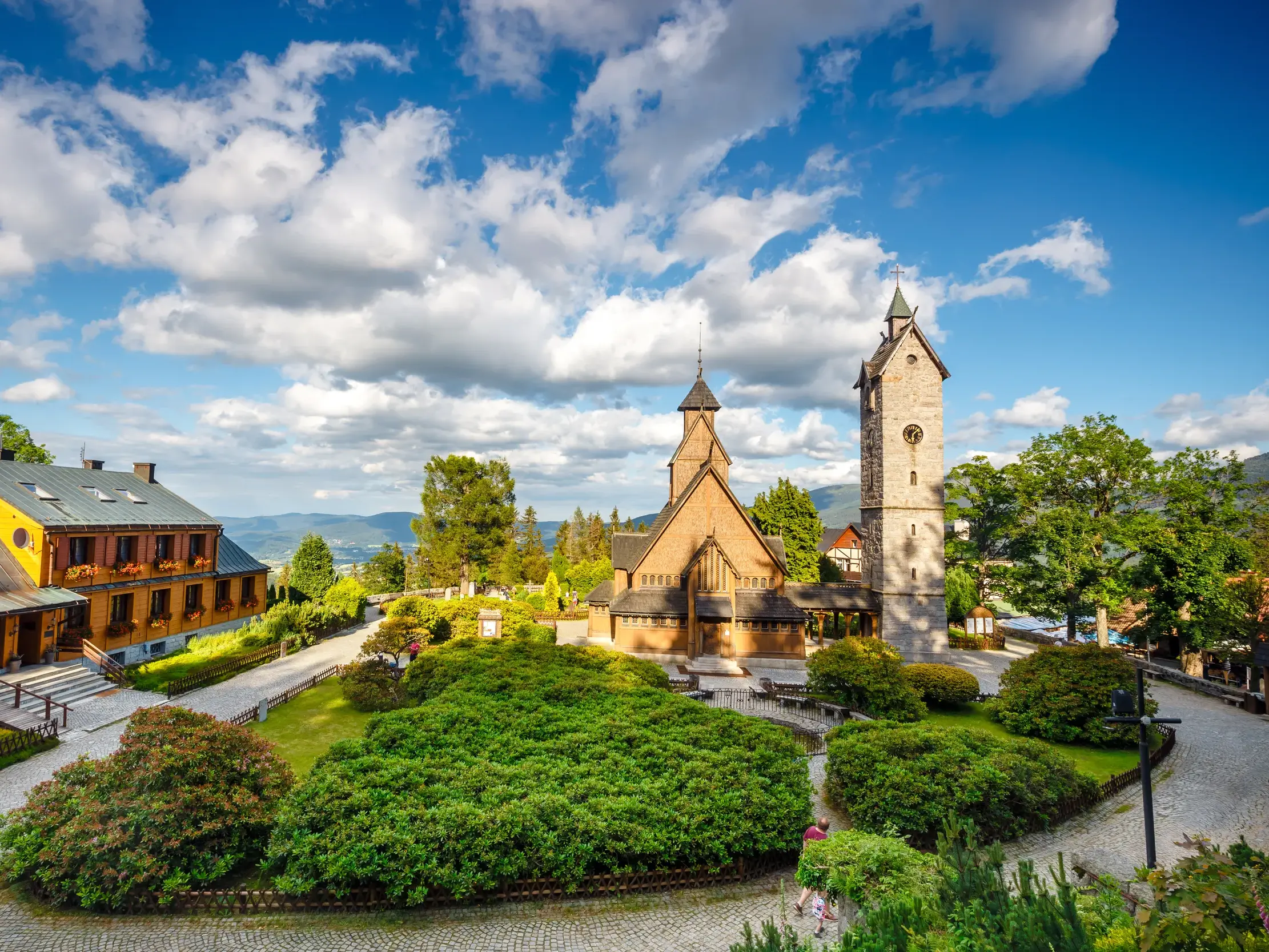 Die hölzerne Stabkirche Wang und ihr Steinturm in Karpacz, umgeben von grünen Gärten vor der Bergkulisse des Riesengebirges.