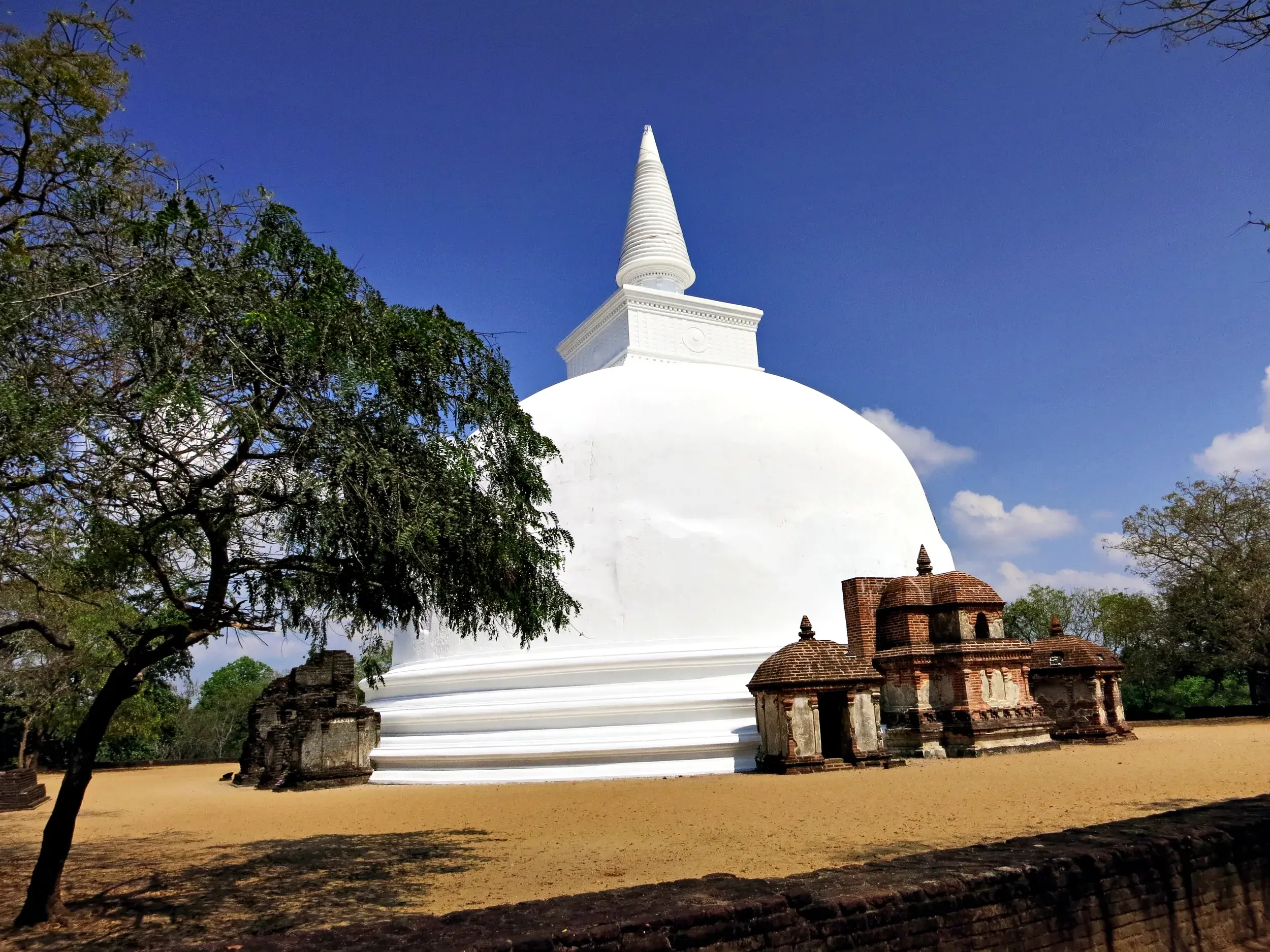 Die strahlend weiße Kiri Vihara Stupa in Polonnaruwa ragt neben alten Ruinen und einem Baum in den blauen Himmel Sri Lankas.