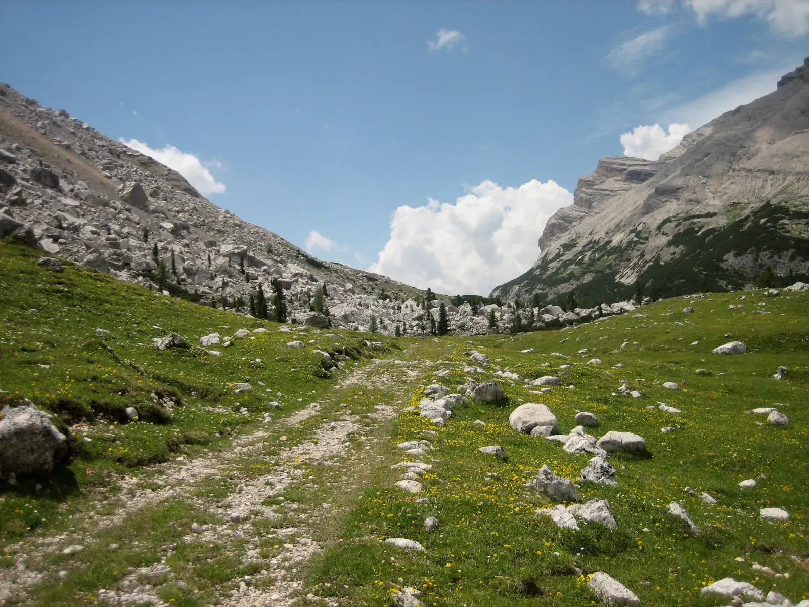 Schotteriger Bergpfad auf einer Almwiese zwischen den steilen, felsigen Gipfeln der Südtiroler Dolomiten bei Sonnenschein.
