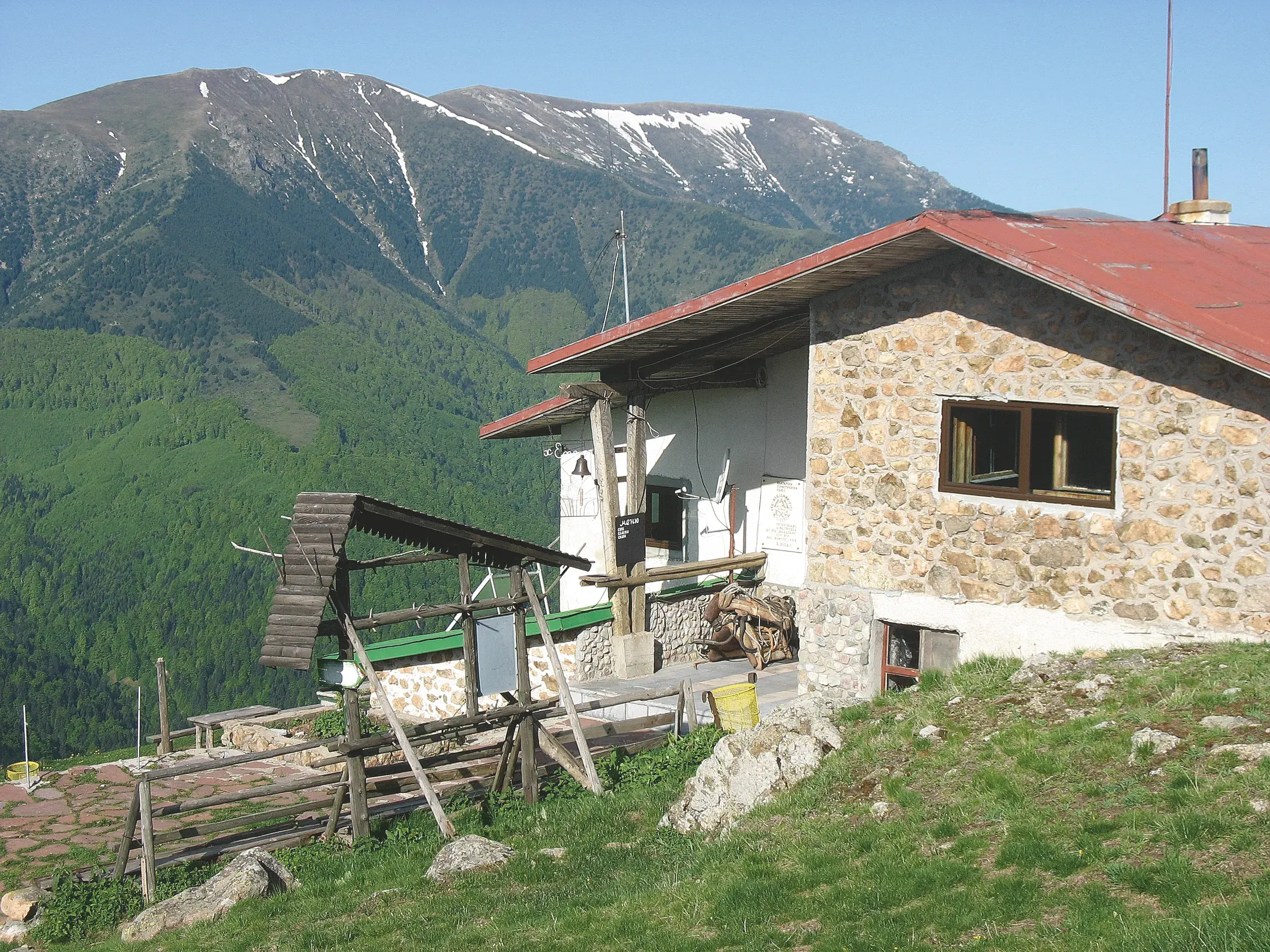 Rustikale Steinhütte mit rotem Dach an einem Grashang vor einem bewaldeten Bergmassiv im Pirin-Gebirge in Bulgarien.