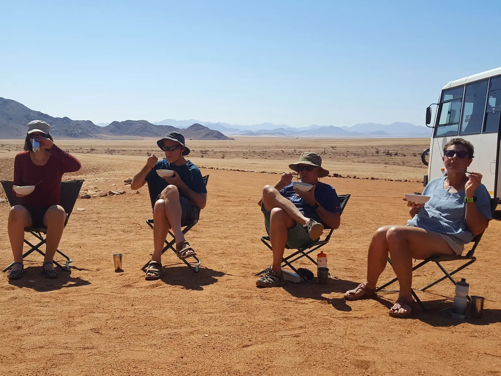 Vier Reisende genießen ihre Mittagspause auf Campingstühlen in der weiten, sonnigen Landschaft der Namib-Wüste in Namibia.