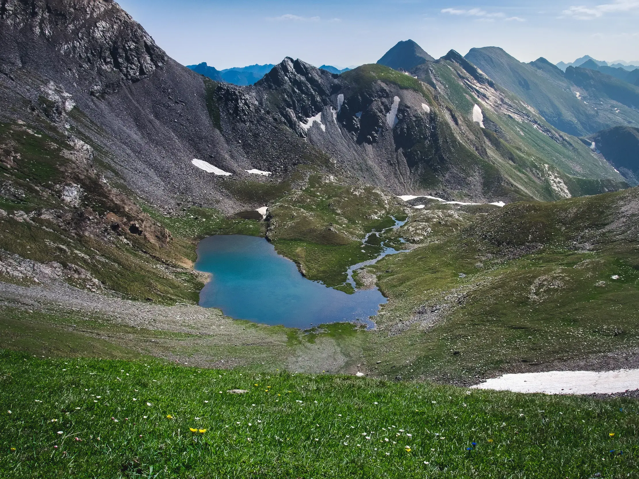 Idyllischer Karsee in den Pyrenäen, umgeben von grünen Almwiesen und schroffen Berggipfeln mit Schneeresten in Andorra.