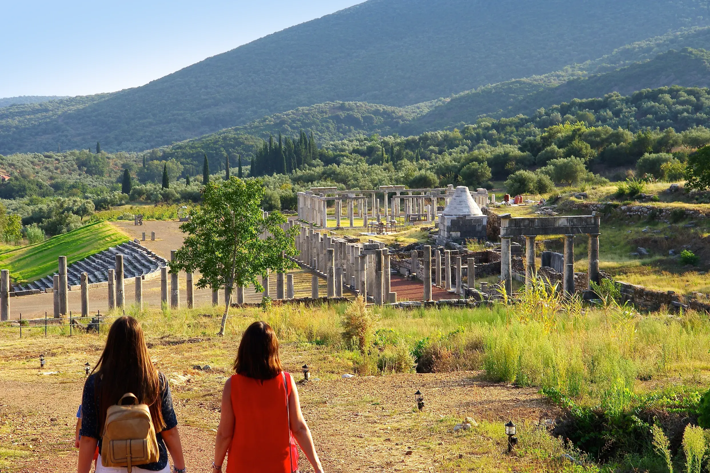 Zwei Frauen erkunden die weitläufige Ausgrabungsstätte Messene mit antiken Säulen und Stadion in der hügeligen Landschaft.