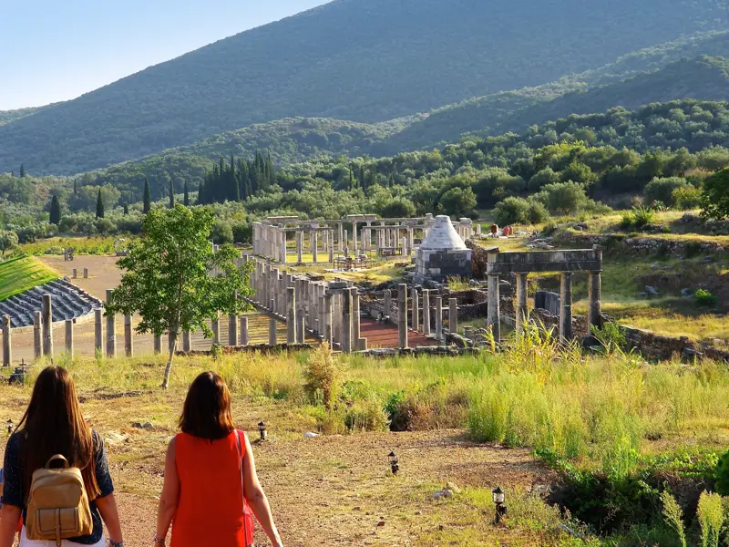 Zwei Frauen erkunden die weitläufige Ausgrabungsstätte Messene mit antiken Säulen und Stadion in der hügeligen Landschaft.