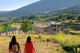 Zwei Frauen erkunden die weitläufige Ausgrabungsstätte Messene mit antiken Säulen und Stadion in der hügeligen Landschaft.