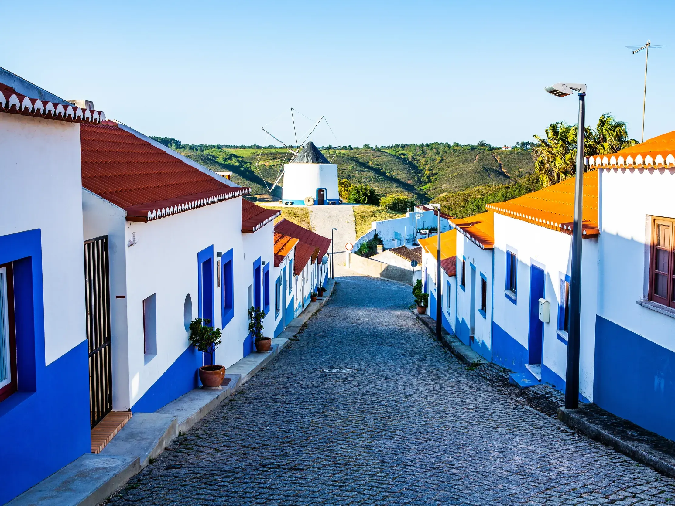 Eine gepflasterte Gasse in Odeceixe, gesäumt von weiß-blauen Häusern, führt bergauf zur historischen Windmühle Portugals.