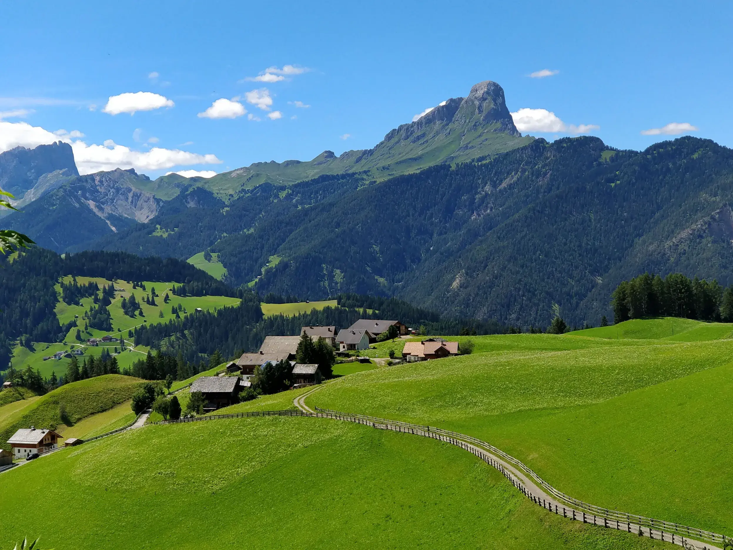 Saftige, grüne Almwiesen und ein kleines Bergdorf bei Wengen vor dem Panorama des Peitlerkofels in den Dolomiten Südtirols.