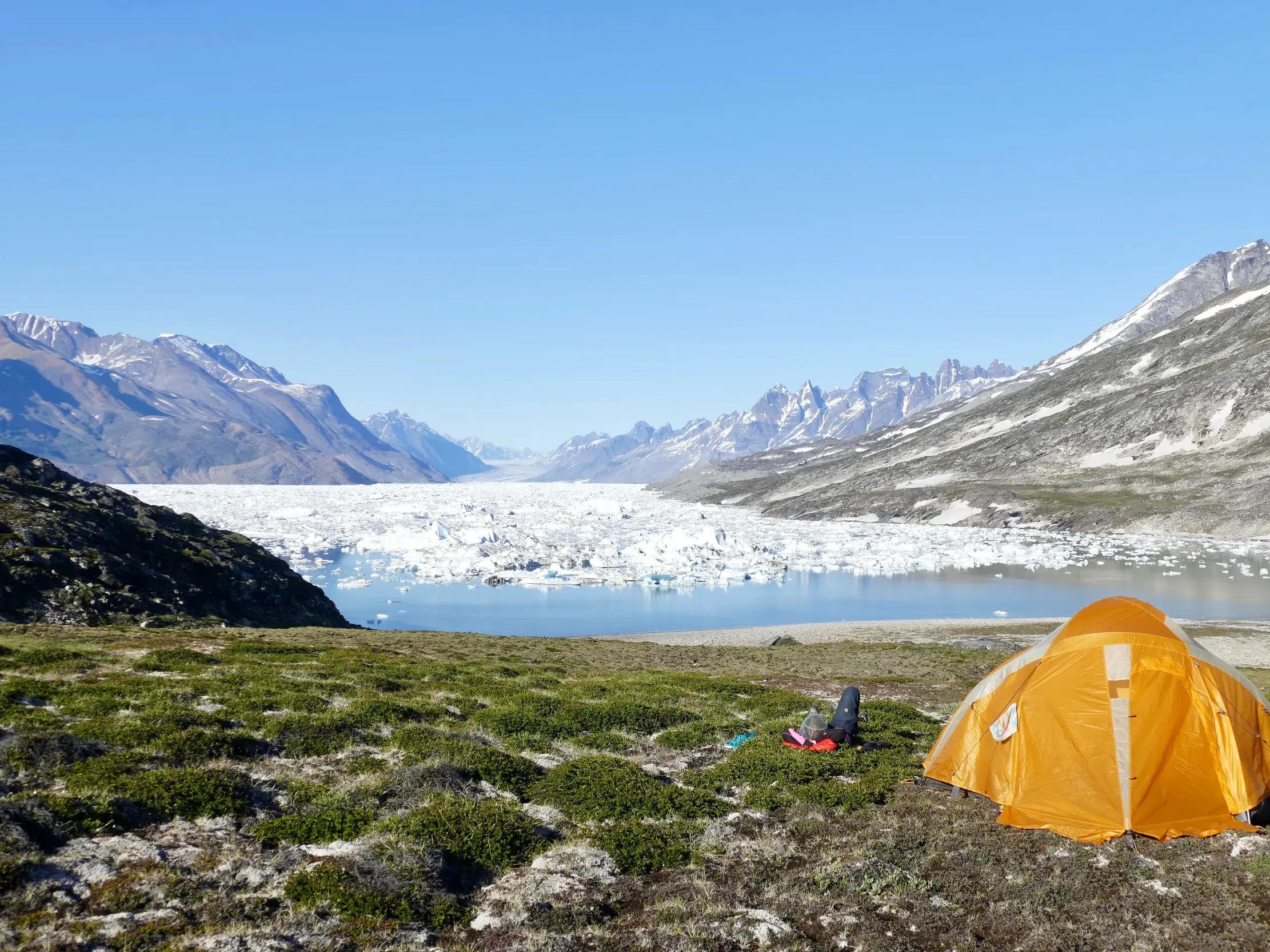 Oranges Zelt auf einer grünen Anhöhe beim Trekking in Grönland mit Panoramablick auf den eisgefüllten Sermilik-Fjord.