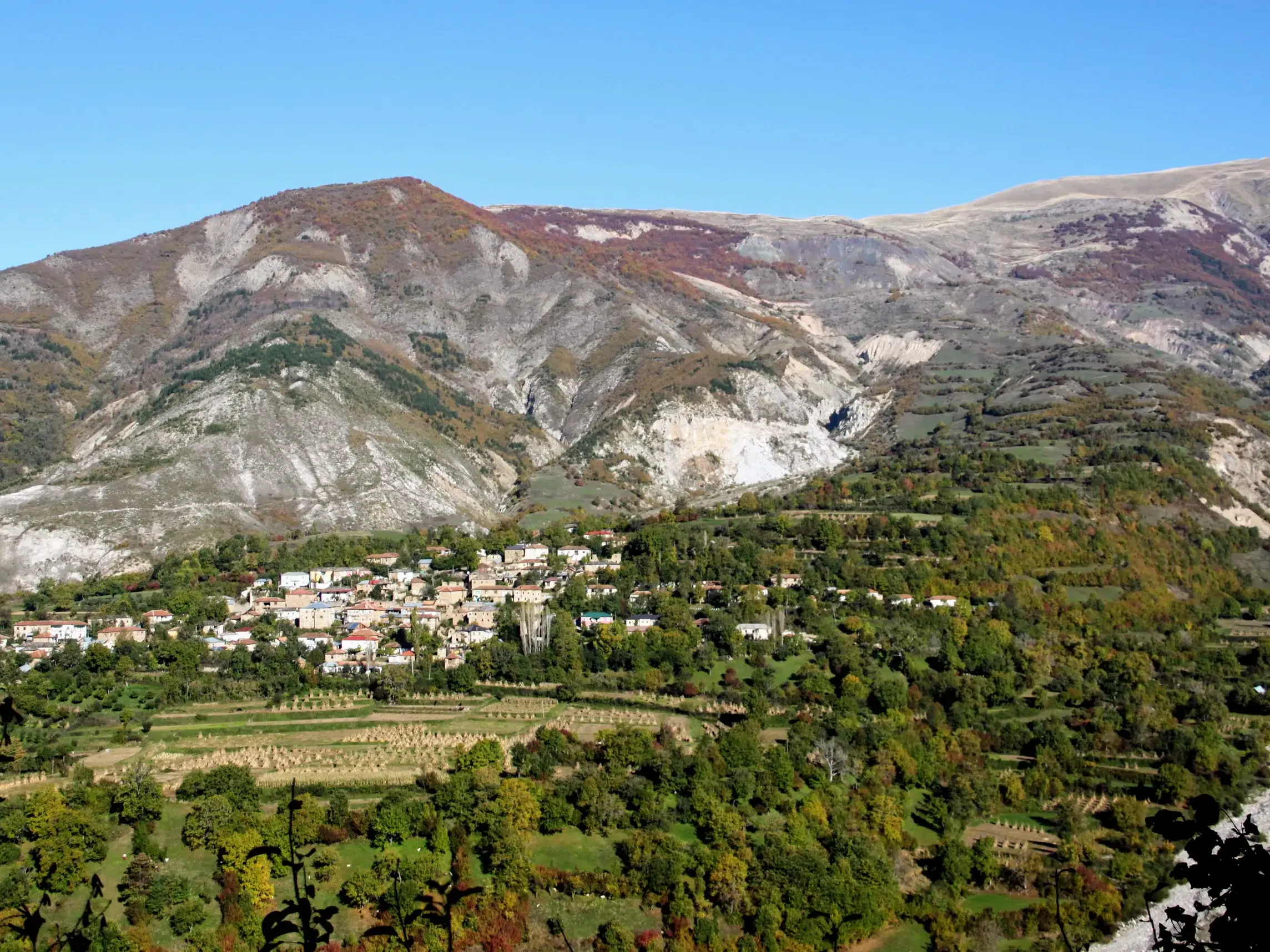 Das albanische Bergdorf Rabdisht liegt an einem sonnigen, grünen Hang mit Terrassenfeldern vor der Kulisse des Korab-Gebirges.