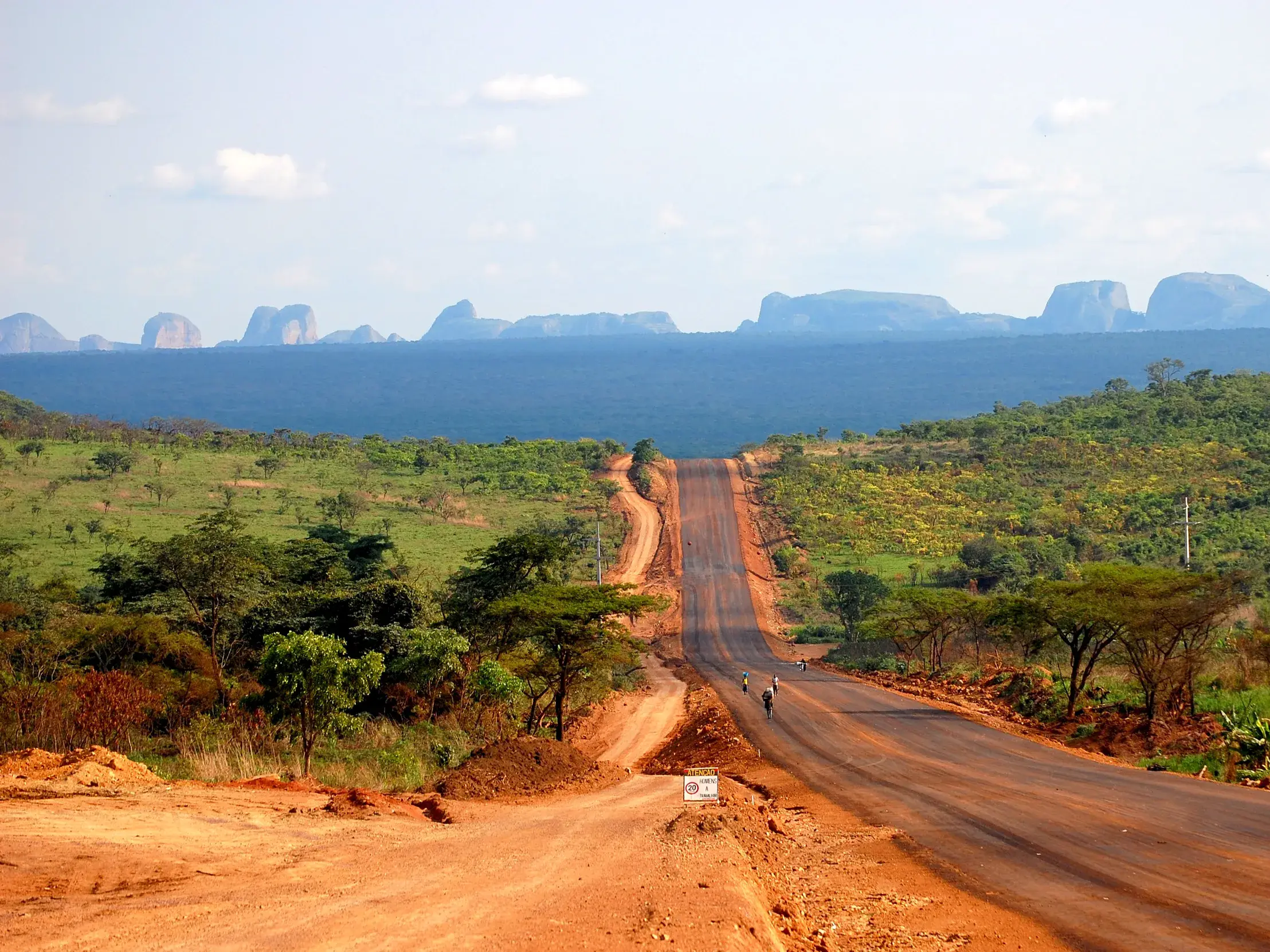 Eine rote Erdstraße führt durch die Savanne Angolas zu den markanten Granitfelsen von Pungo Andongo am Horizont.
