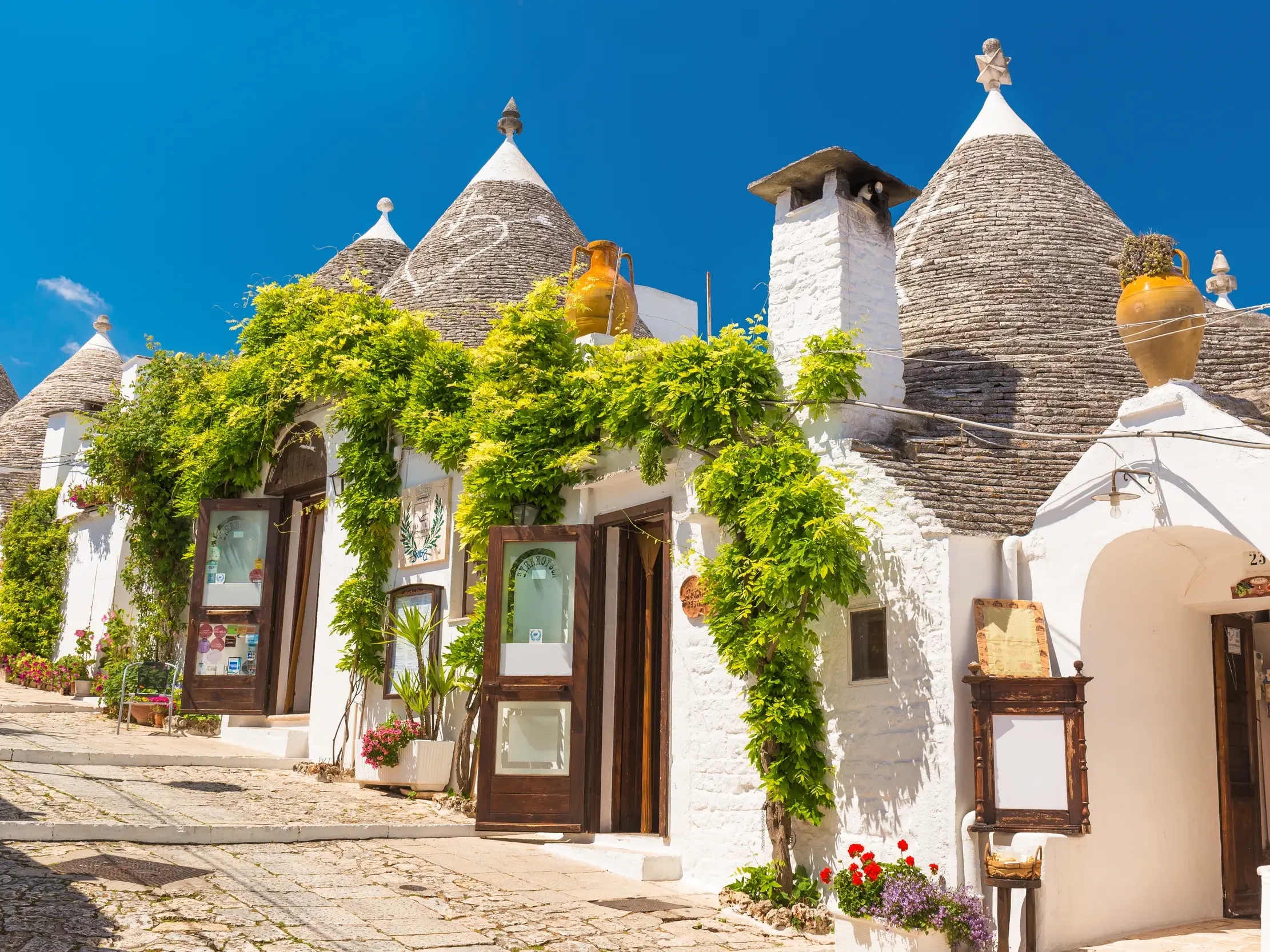 Sonnige Gasse in Alberobello mit weißen Trulli-Rundsteinbauten, deren Fassaden mit grünen Rankpflanzen bewachsen sind.