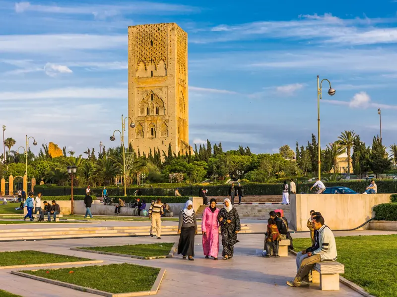 Drei Frauen in traditioneller Kleidung spazieren an einem sonnigen Tag durch den Park vor dem historischen Hassan-Turm in Rabat.