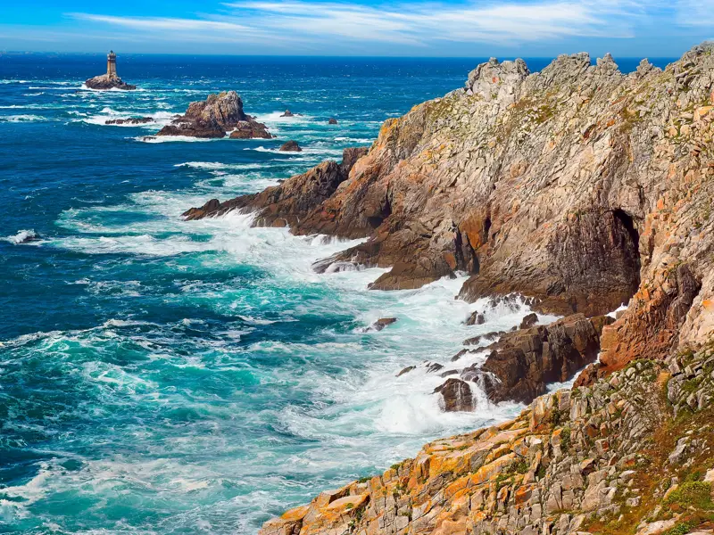 Die wilde Felsküste der Pointe du Raz in der Bretagne. Wellen branden an die Klippen, Leuchttürme am Horizont.