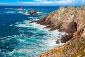 Die wilde Felsküste der Pointe du Raz in der Bretagne. Wellen branden an die Klippen, Leuchttürme am Horizont.
