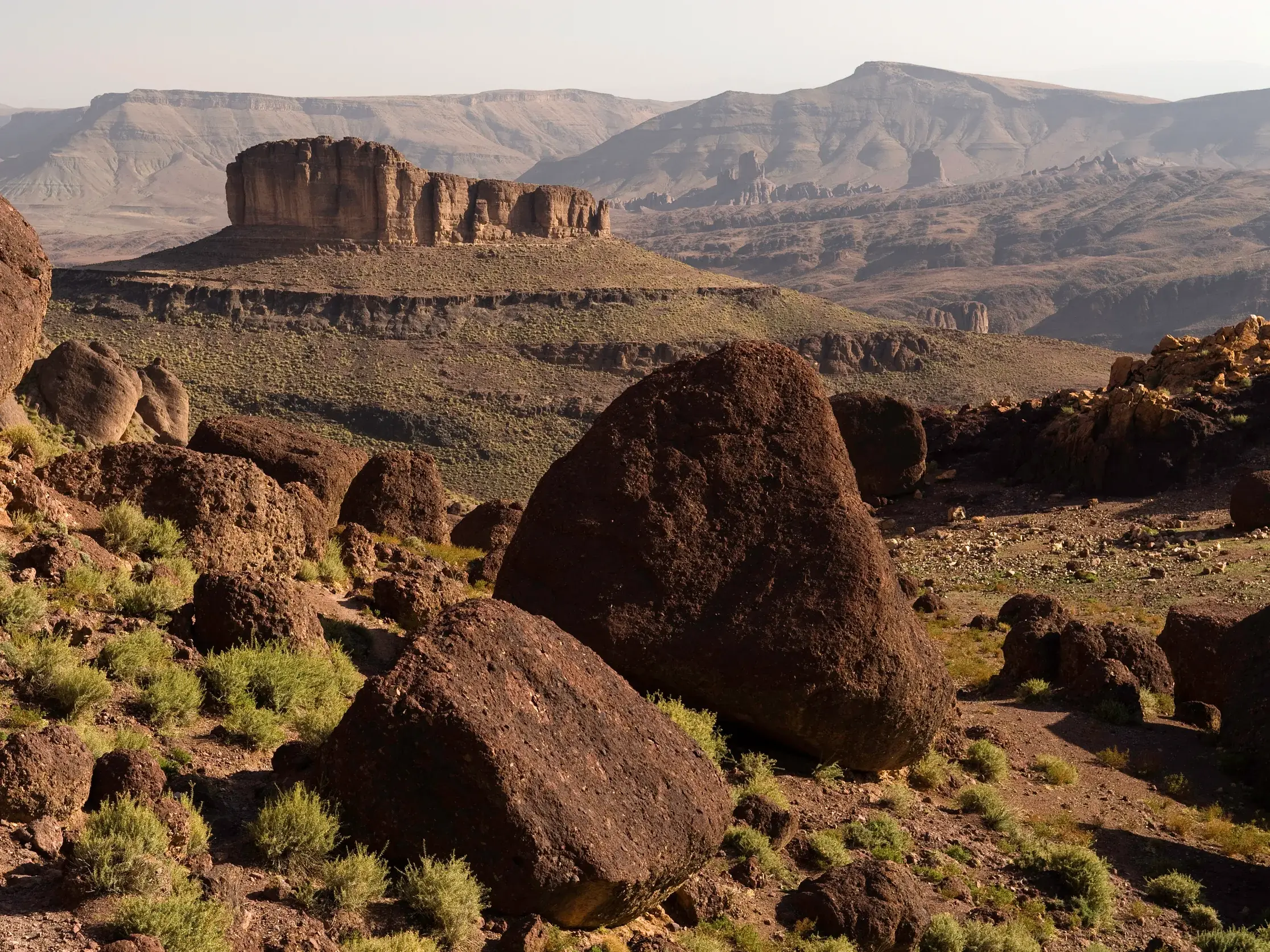 Weite Vulkanlandschaft im Saghro-Gebirge Marokkos mit dem markanten Tafelberg Bab n'Ali und Felsbrocken im Vordergrund.
