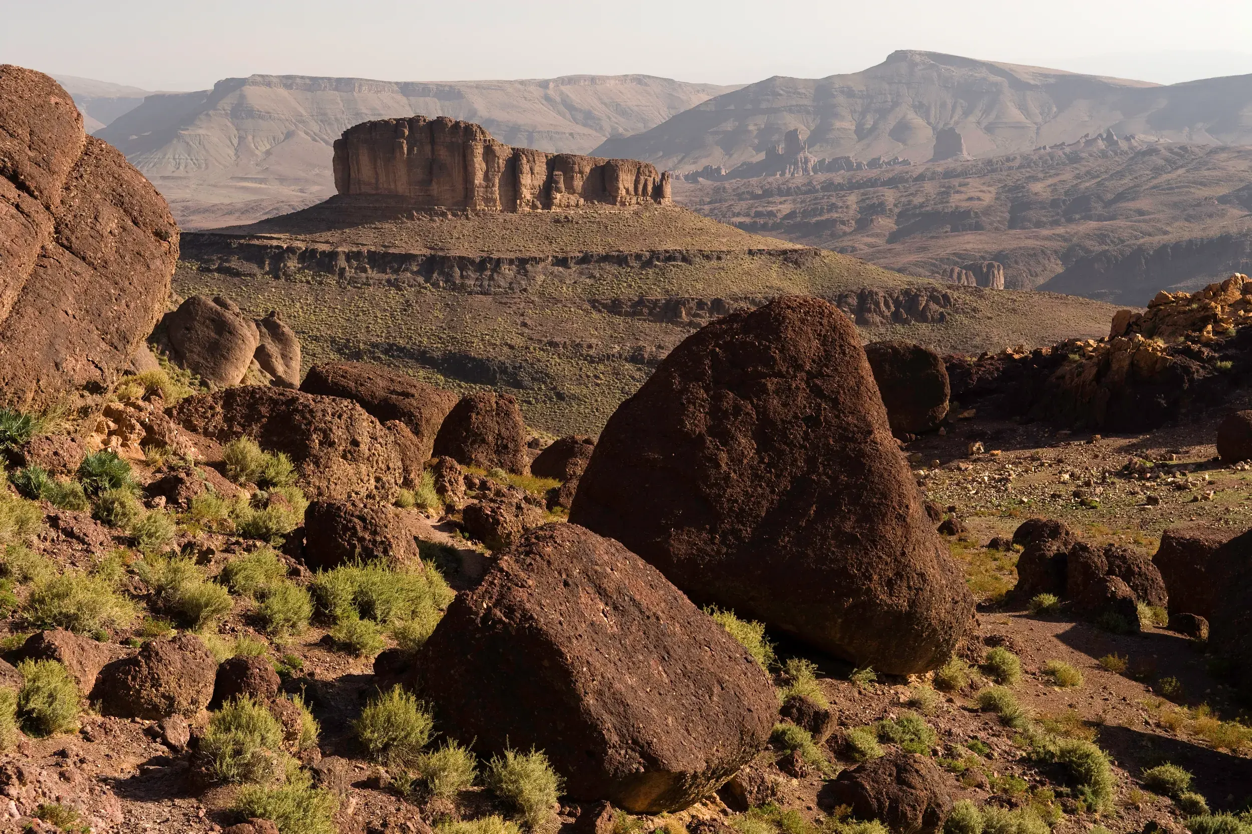 Weite Vulkanlandschaft im Saghro-Gebirge Marokkos mit dem markanten Tafelberg Bab n'Ali und Felsbrocken im Vordergrund.