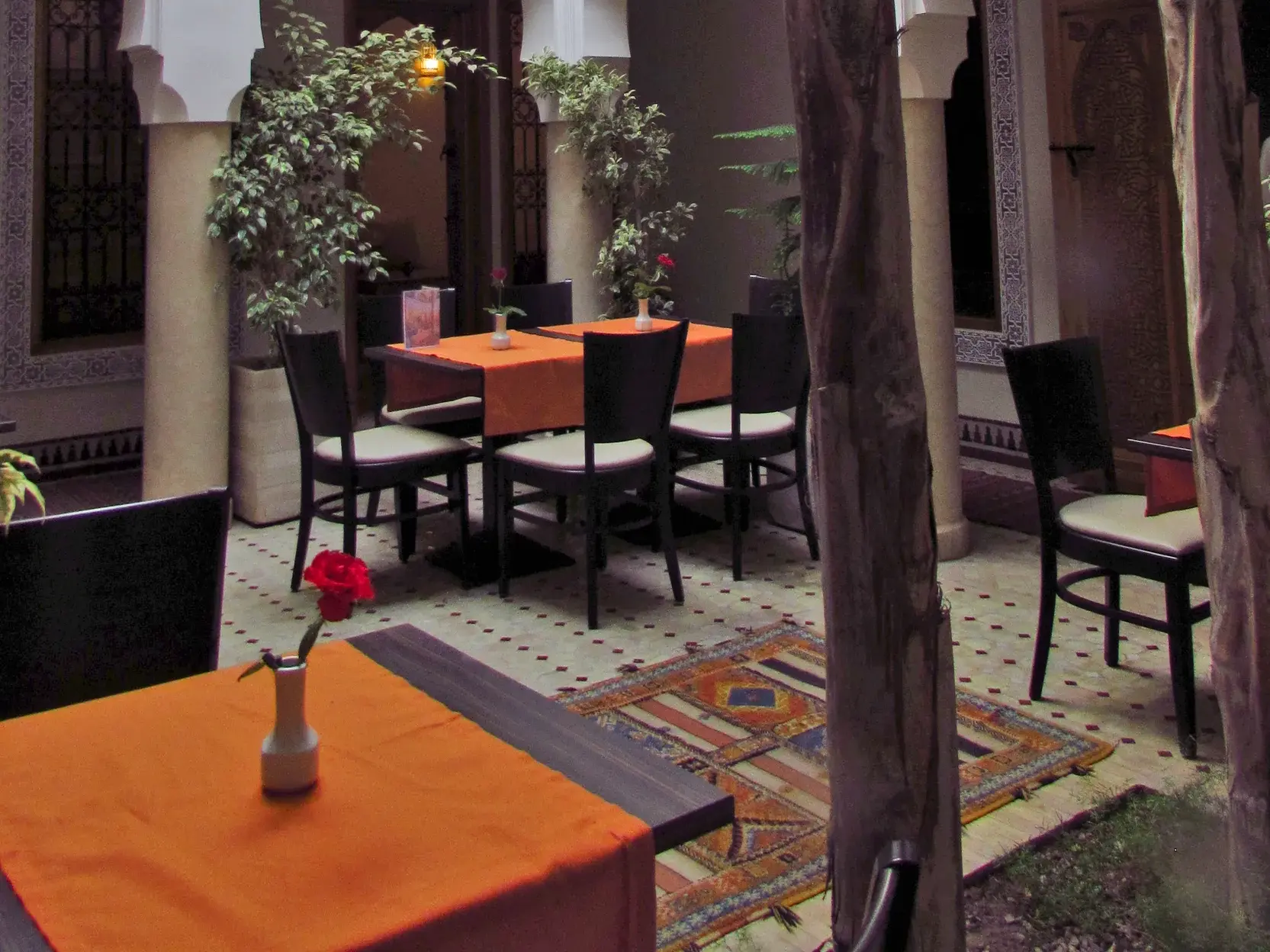 Gedeckte Tische mit orangefarbenen Decken im ruhigen, bepflanzten Innenhof eines traditionellen Riads in Marokko.