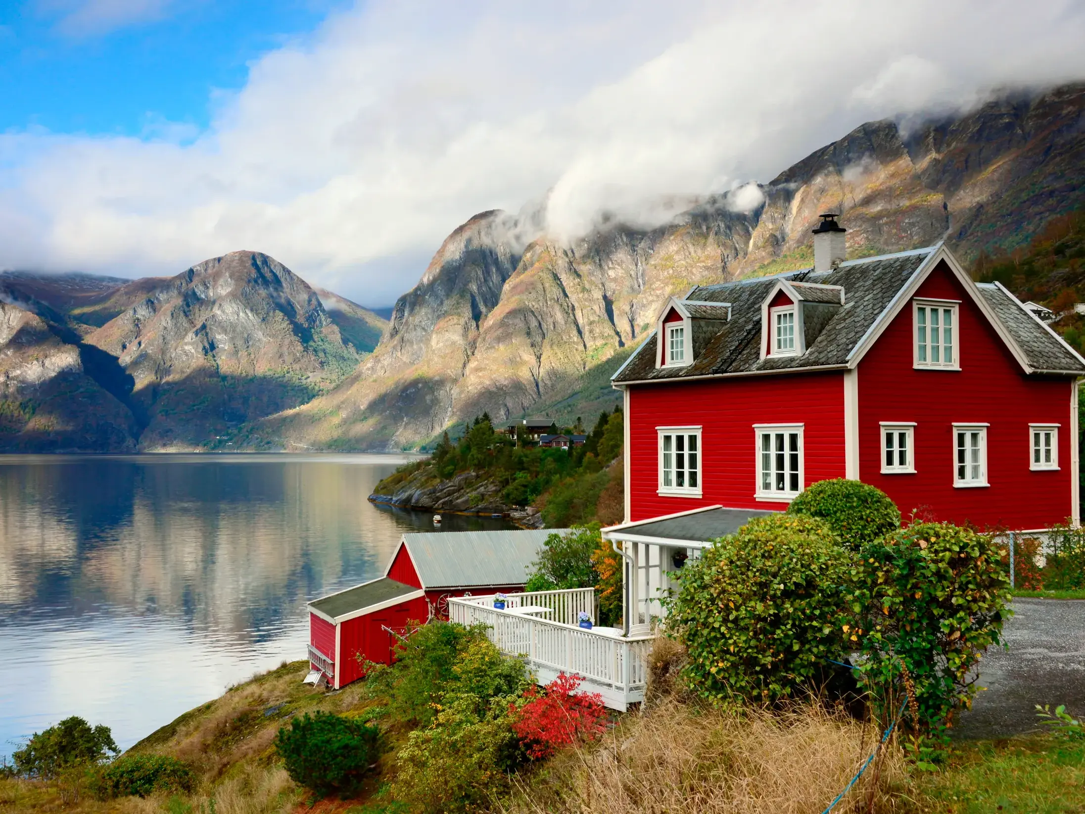 Malerisches rotes Holzhaus an einem norwegischen Fjord, dessen ruhiges Wasser die wolkenverhangenen Berge reflektiert.