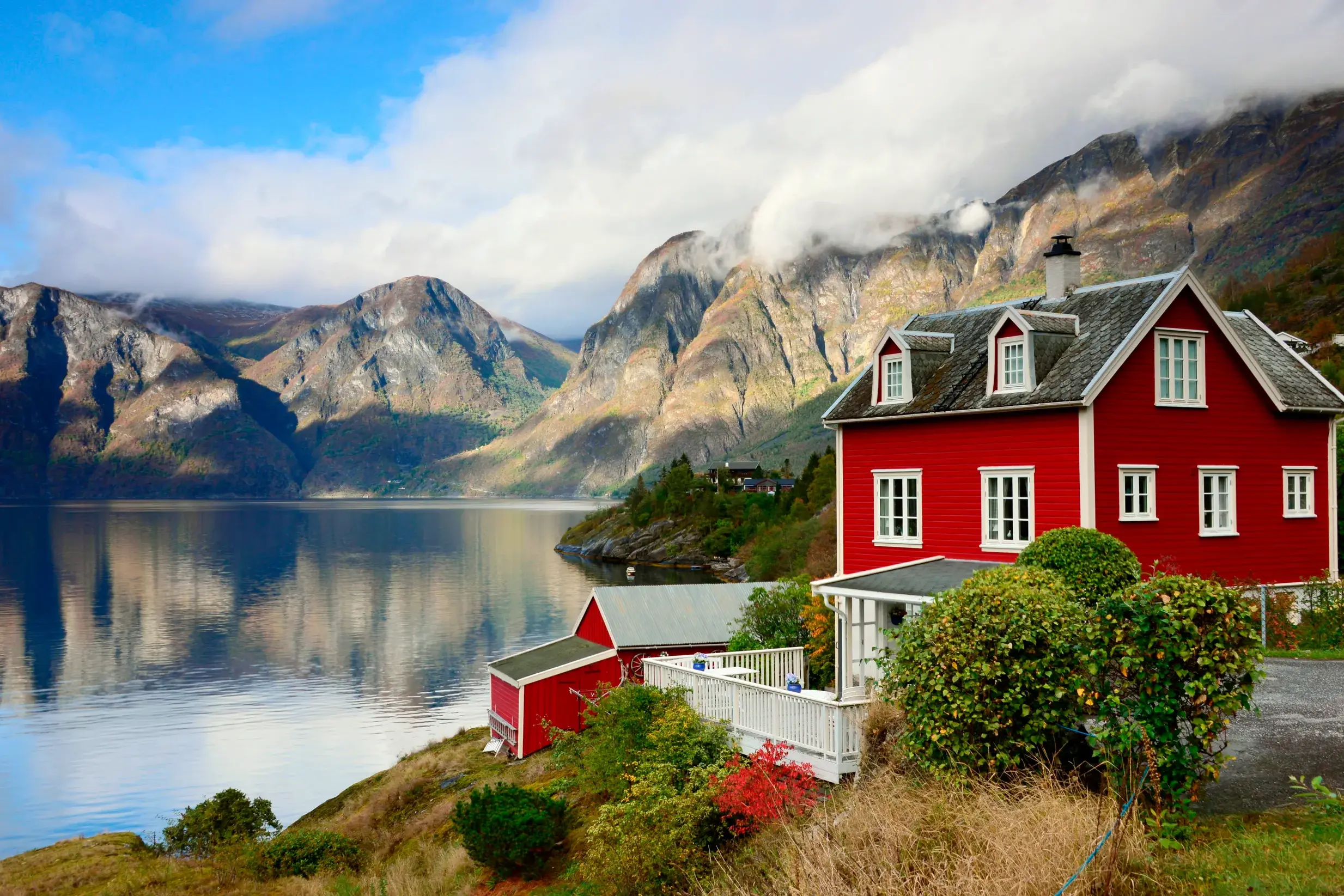 Malerisches rotes Holzhaus an einem norwegischen Fjord, dessen ruhiges Wasser die wolkenverhangenen Berge reflektiert.