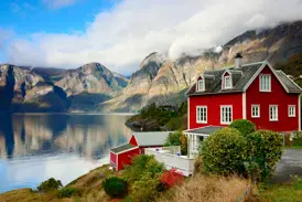 Malerisches rotes Holzhaus an einem norwegischen Fjord, dessen ruhiges Wasser die wolkenverhangenen Berge reflektiert.