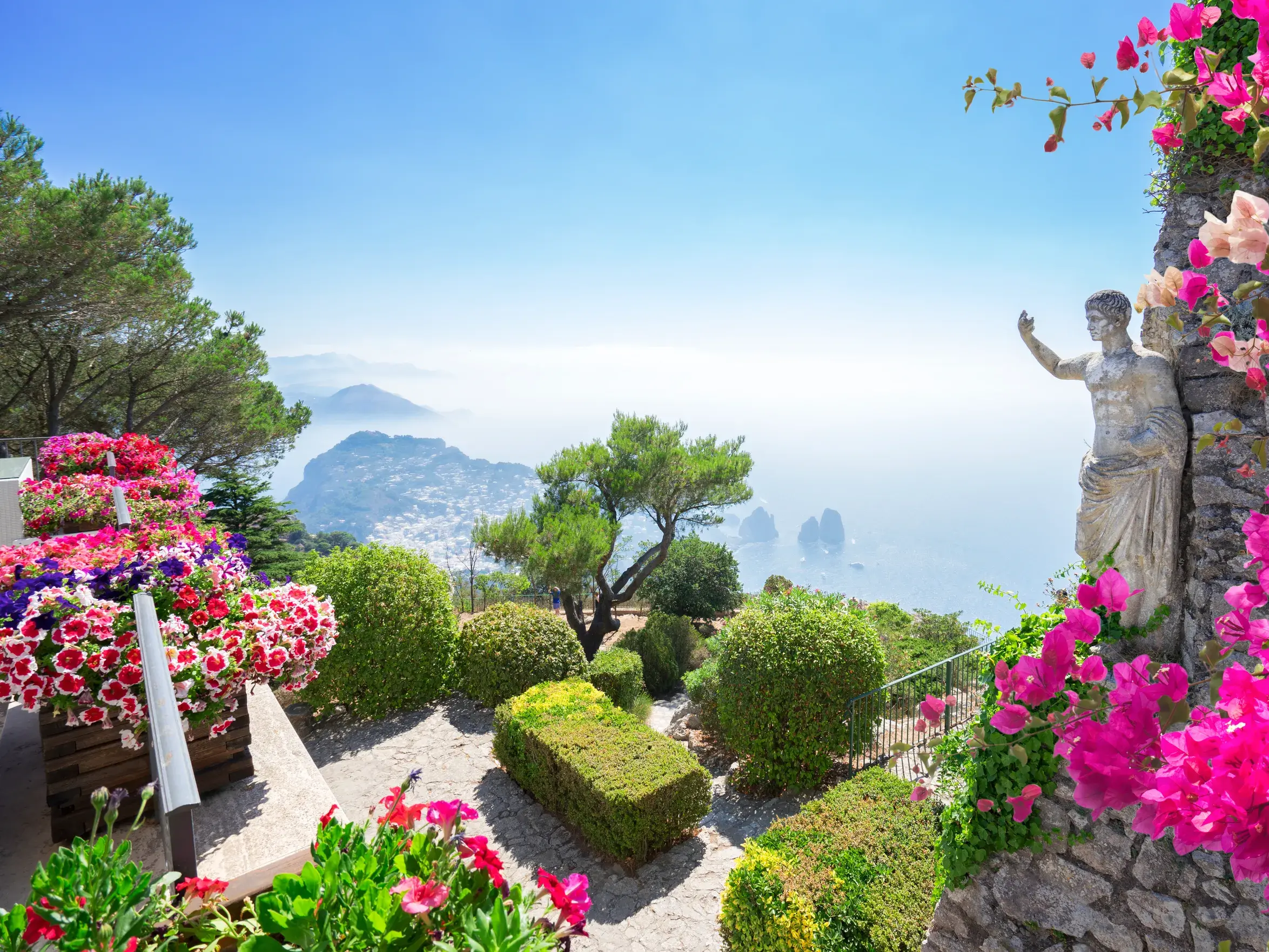 Von einem blühenden Garten mit Statue auf Capri eröffnet sich ein Panoramablick über das tiefblaue Meer und die Küste.