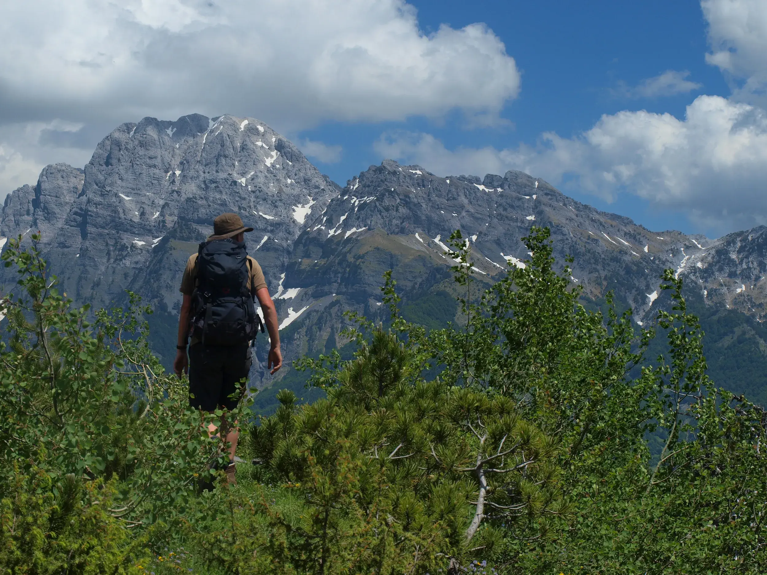 Ein Wanderer mit Trekkingrucksack und Hut genießt auf einem grünen Pfad die Aussicht auf die schroffen Gipfel im Valbona-Tal.
