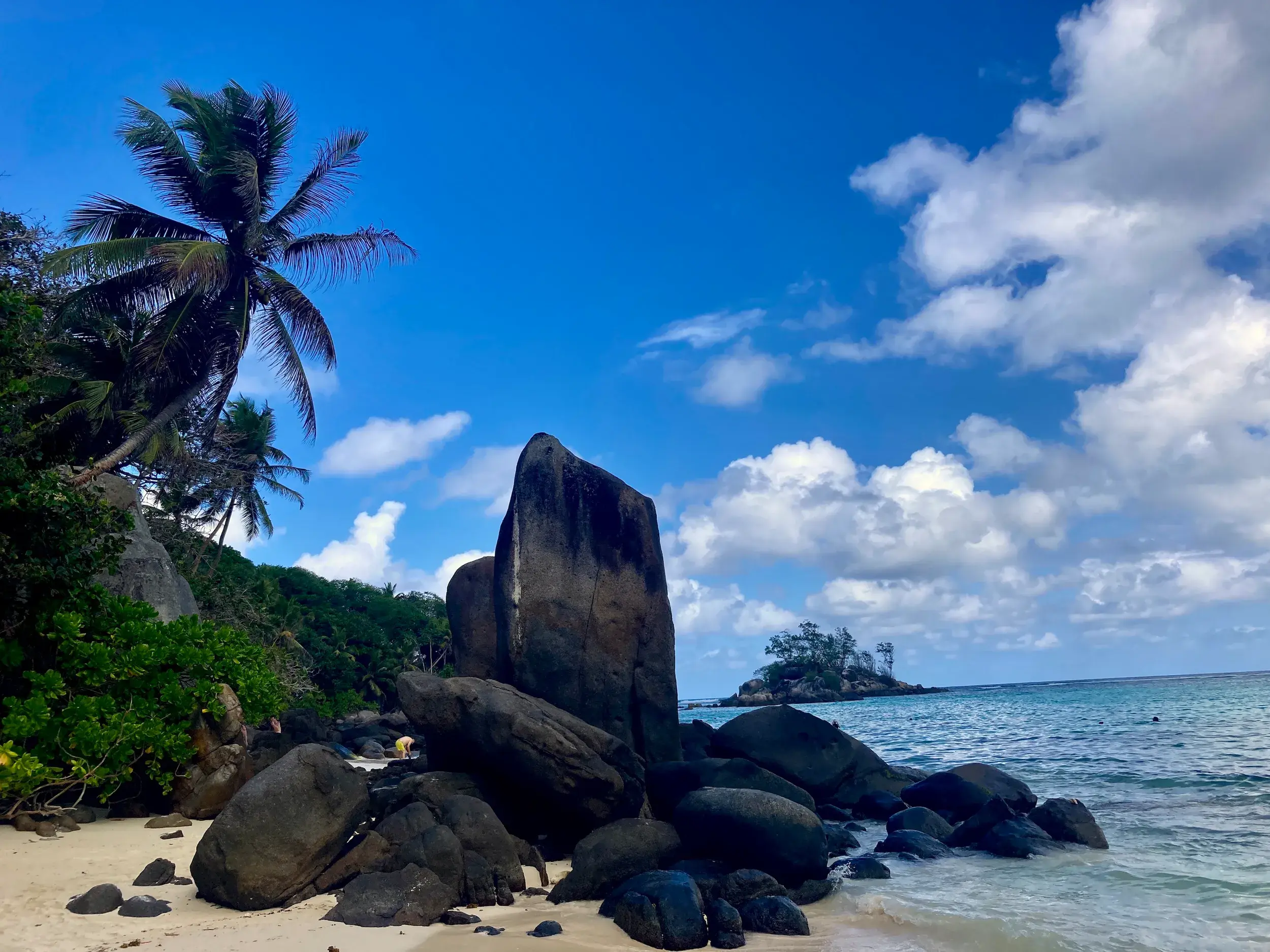 Imposante Granitfelsen an einem Sandstrand auf den Seychellen mit Palmen und Blick auf eine kleine Insel im Indischen Ozean.