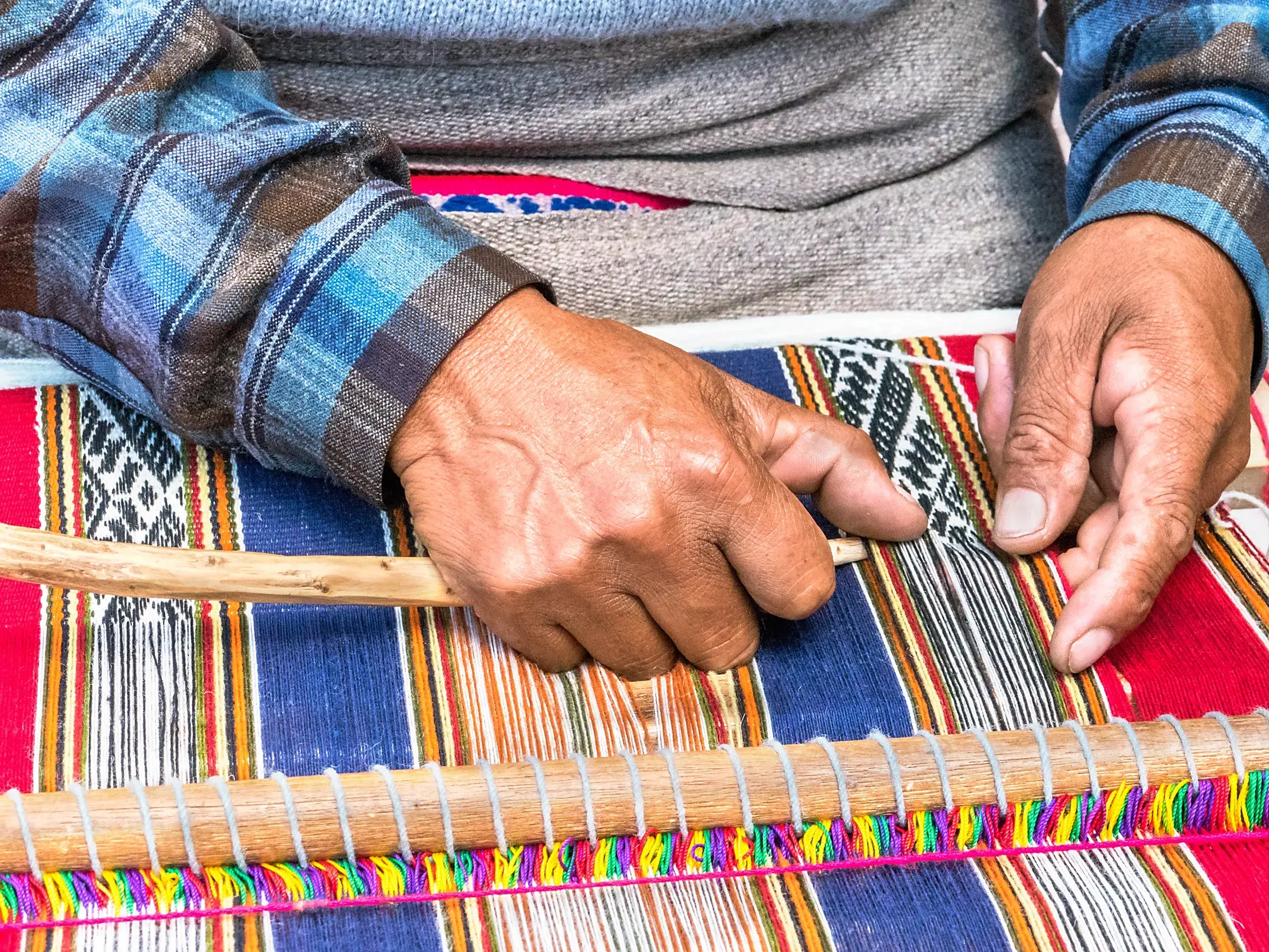 Nahaufnahme der Hände eines Webers, der in Peru an einem Handwebstuhl einen farbenfrohen Stoff mit traditionellen Mustern webt.