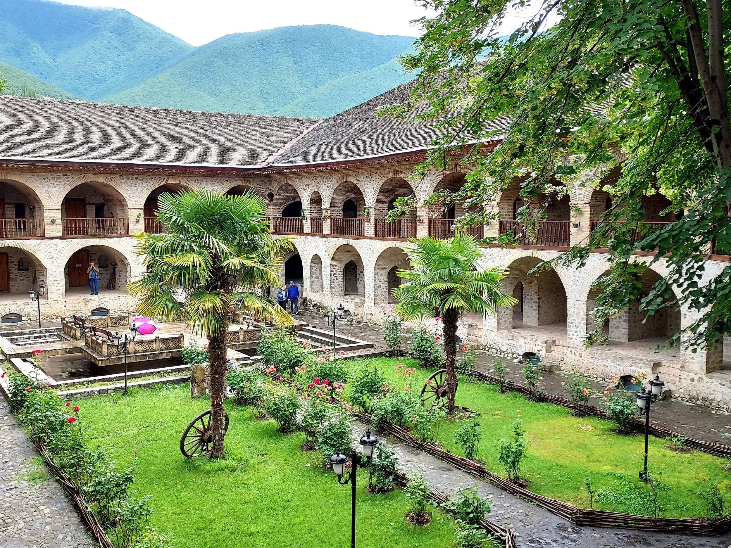 Grüner Innenhof der historischen Karawanserei in Sheki, Aserbaidschan, mit steinernen Arkaden, Palmen und Bergblick.
