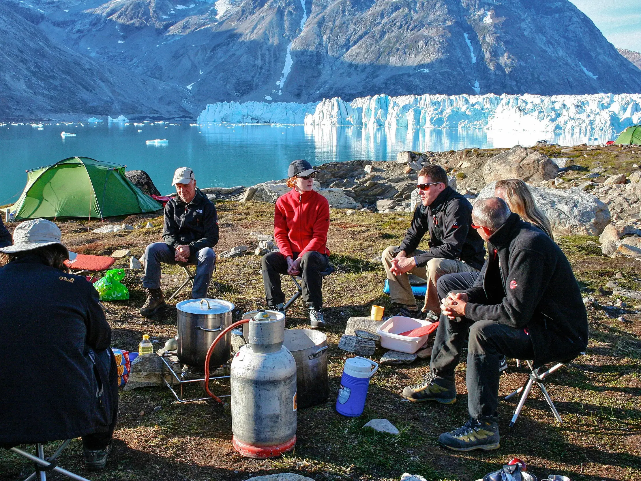 Eine Hauser-Trekkinggruppe beim gemeinsamen Kochen im Zeltcamp am Ufer eines Fjords in Ostgrönland vor einer Gletscherwand.