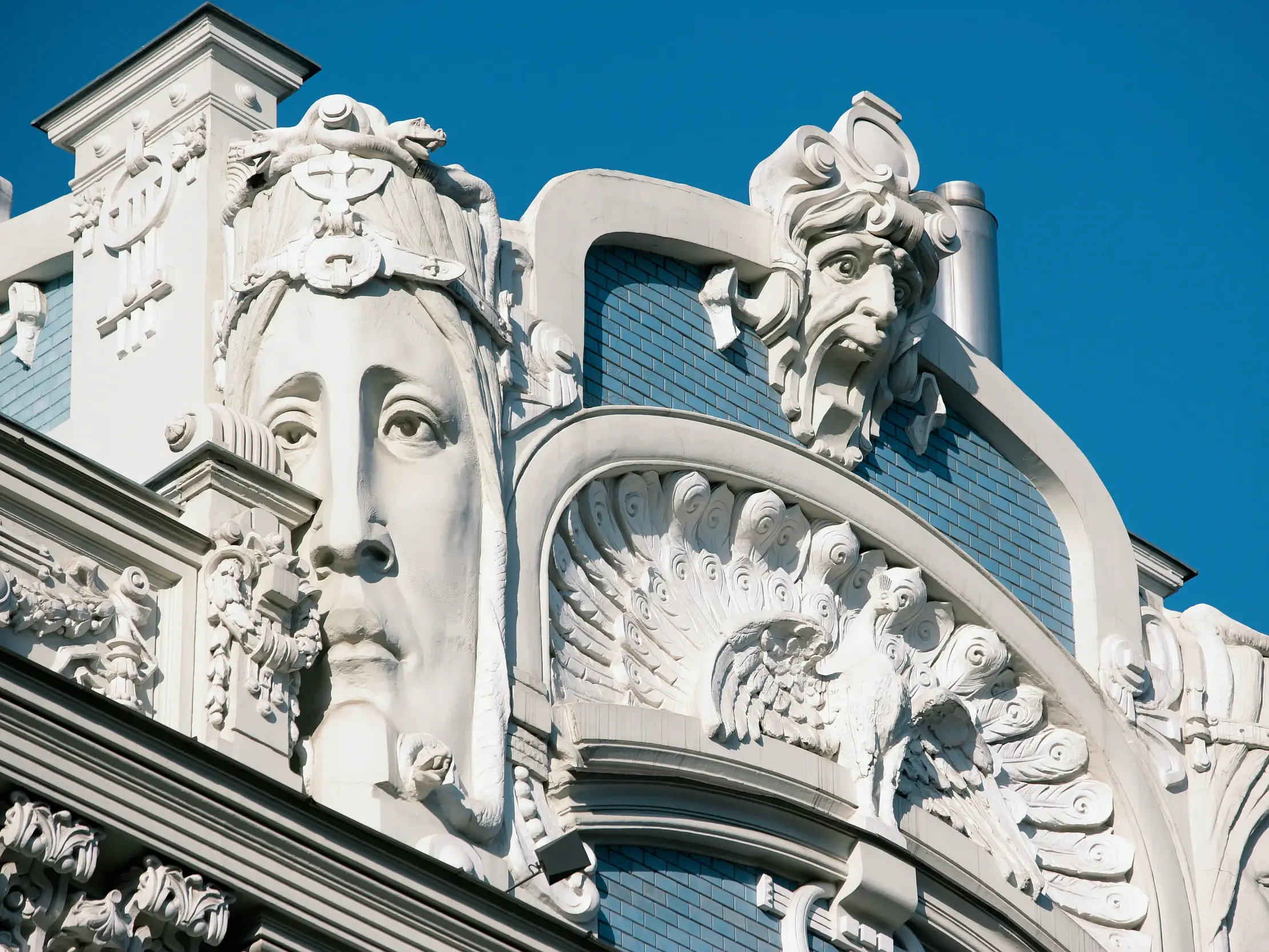 Nahaufnahme der prunkvollen Jugendstil-Fassade eines Hauses in Riga mit weißen Skulpturen von Gesichtern und einem Pfau.
