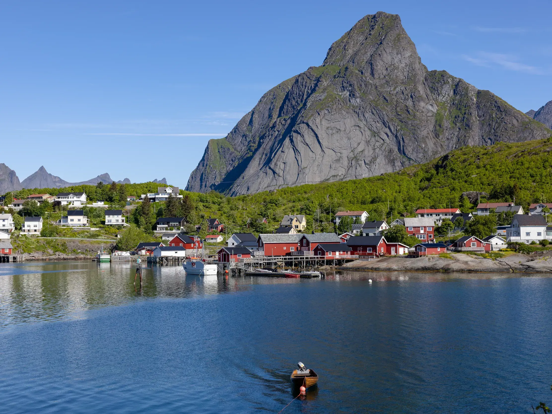 Idyllisches Fischerdorf mit roten Holzhäusern am Wasser auf den Lofoten entlang der Postschiffroute der Hurtigruten, überragt von einem markanten Felsberg bei Sonnenschein.