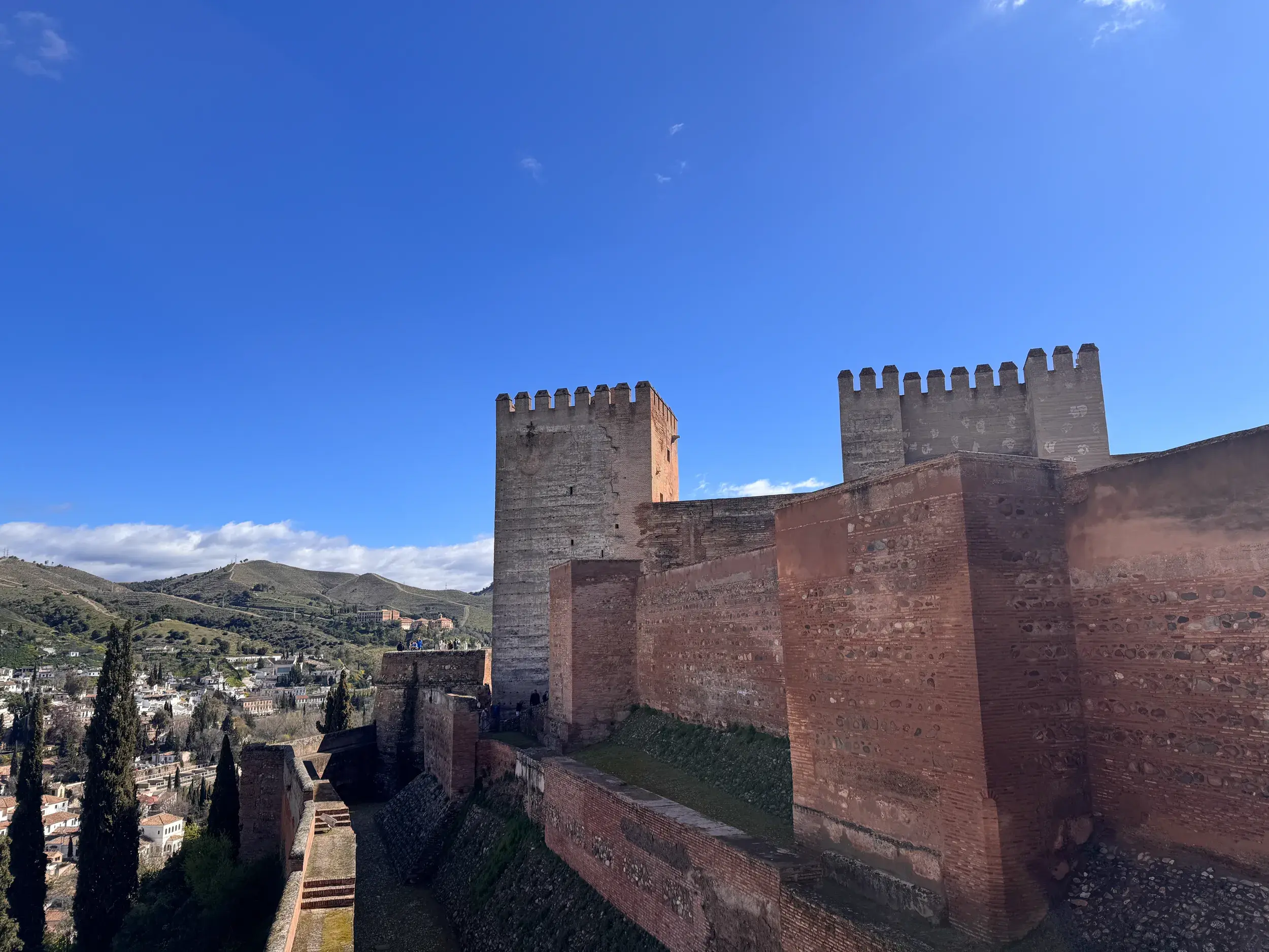 Die Wehrmauern und Türme der maurischen Festung Alhambra in Granada unter blauem Himmel mit Blick auf die Hügel Andalusiens.