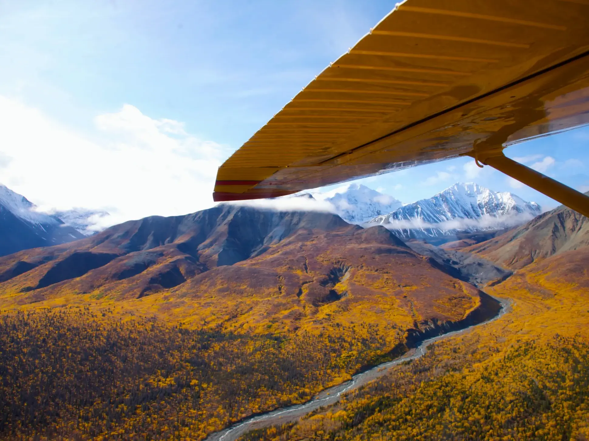 Gelbe Tragfläche eines Kleinflugzeugs über herbstlicher Flusslandschaft und den Bergen des Yukon in Kanada.