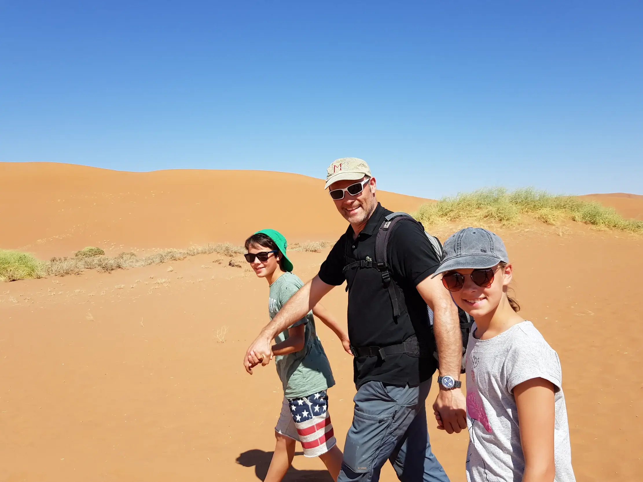Ein Vater wandert mit seinem Sohn und seiner Tochter durch die leuchtend roten Sanddünen der Namib-Wüste in Namibia.