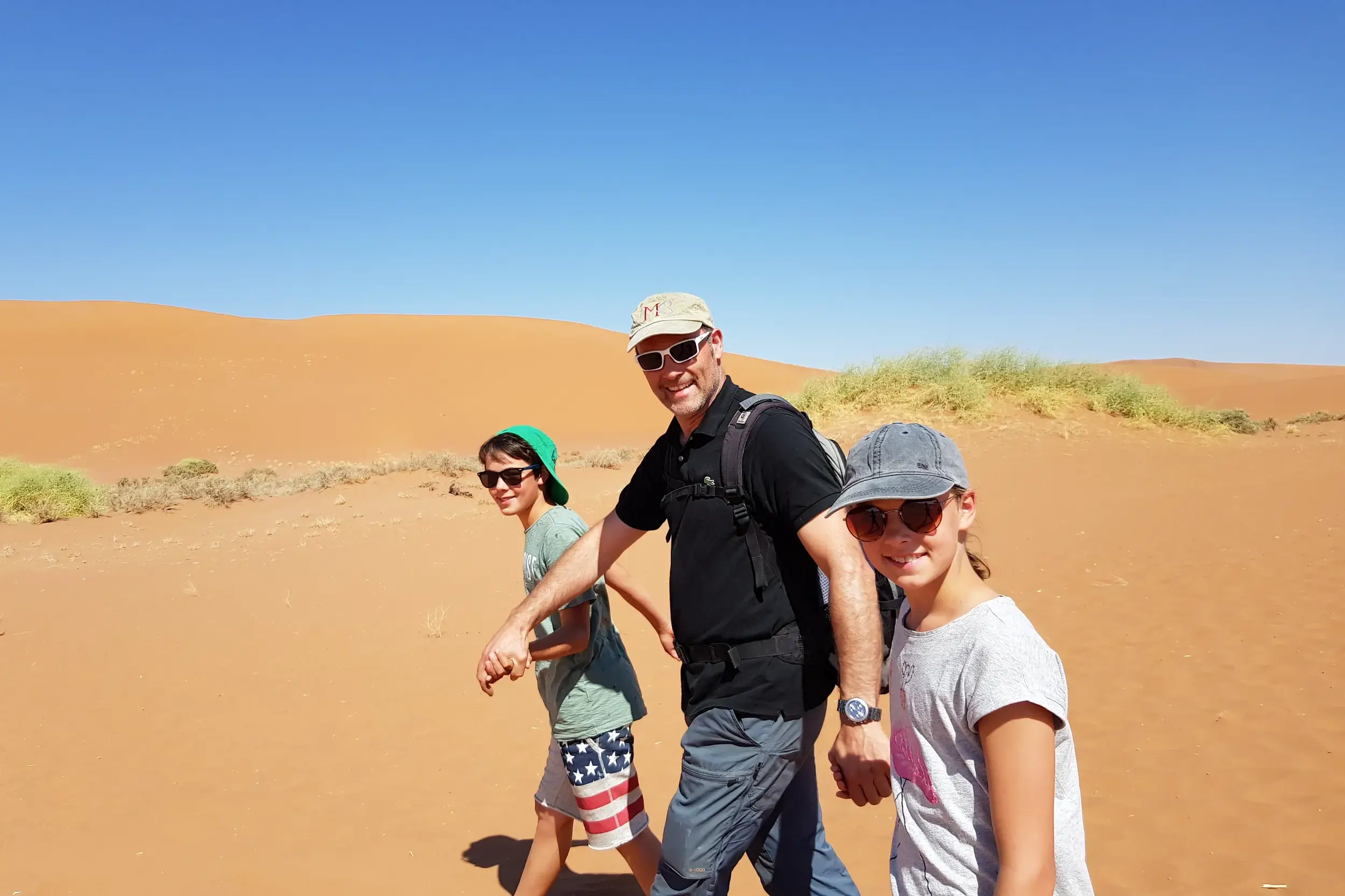 Ein Vater wandert mit seinem Sohn und seiner Tochter durch die leuchtend roten Sanddünen der Namib-Wüste in Namibia.