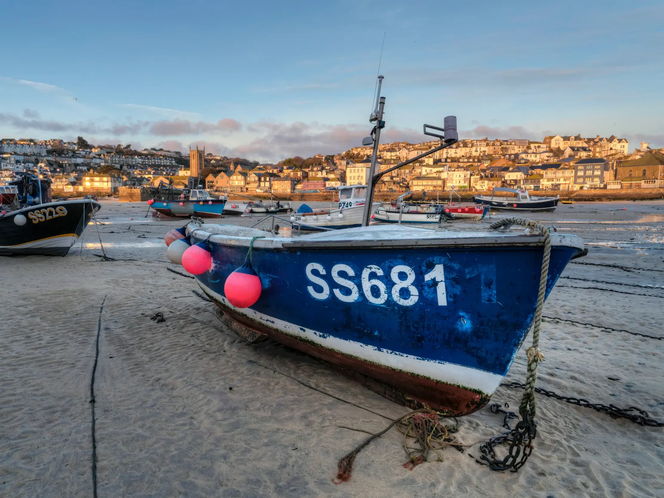 Blaues Fischerboot liegt bei Ebbe auf dem sandigen Grund des Hafens von St Ives. Die Stadt leuchtet im Morgenlicht.
