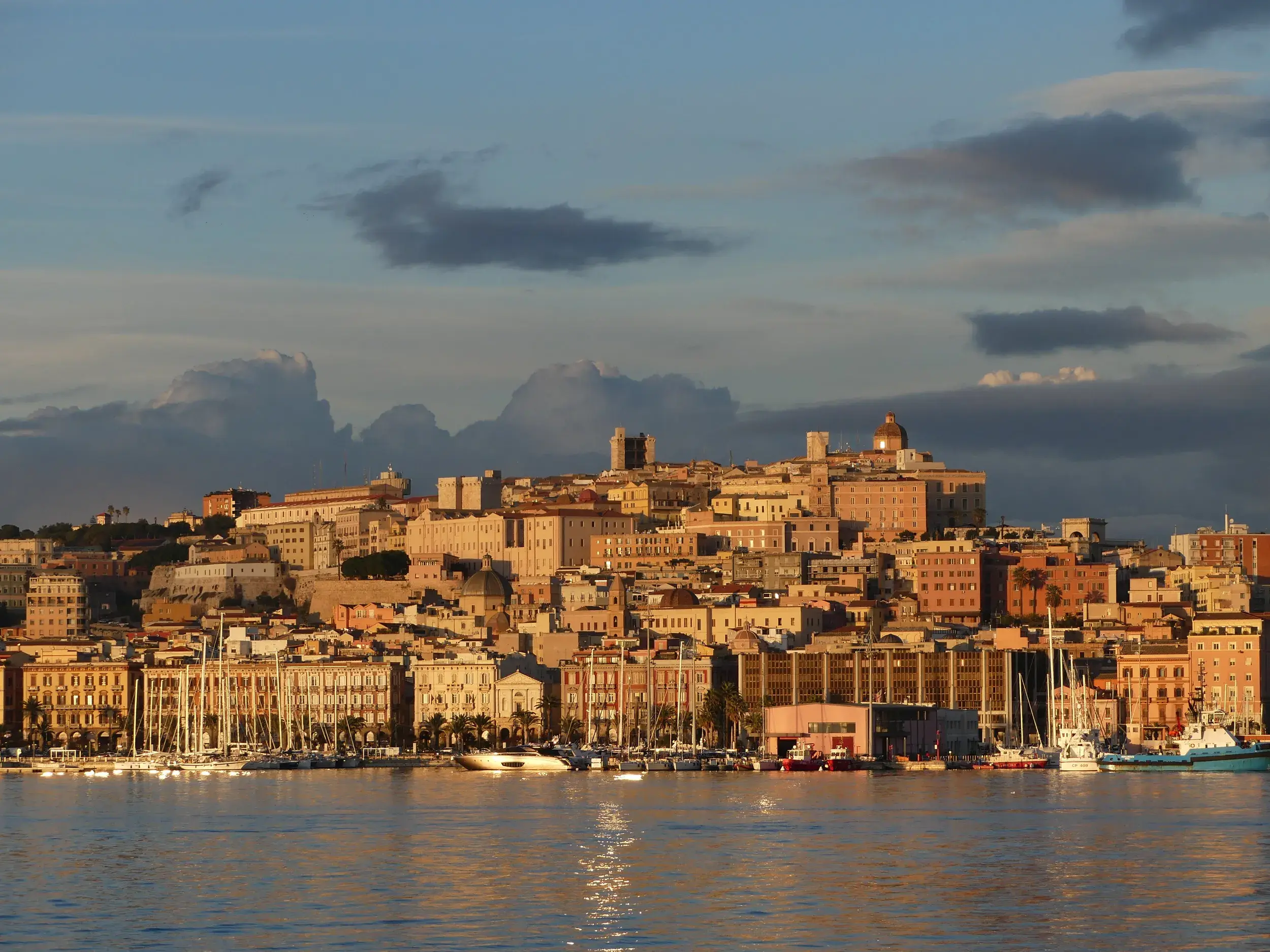 Die historische Altstadt von Cagliari auf Sardinien, im warmen Abendlicht vom Hafen aus betrachtet, mit Segelbooten im Vordergrund.