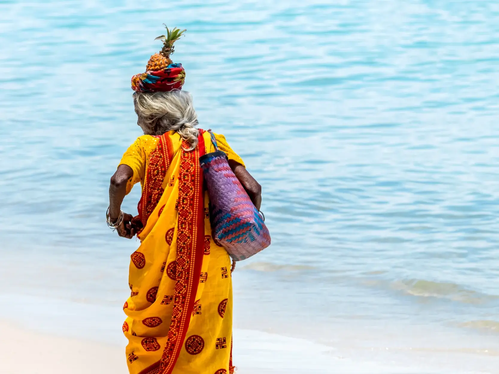 Eine einheimische Frau in gelbem Sari balanciert eine Ananas auf ihrem Kopf und spaziert am türkisen Meer von Mauritius entlang.